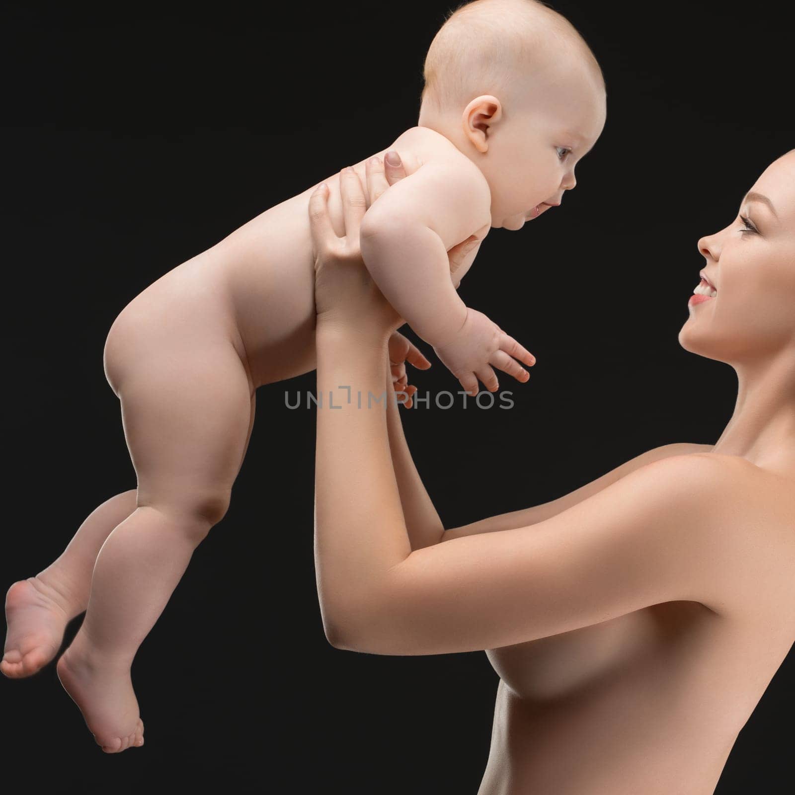A mother joyfully lifts her baby while both share a playful and loving expression, showcasing a bond in a serene setting with a dark background.