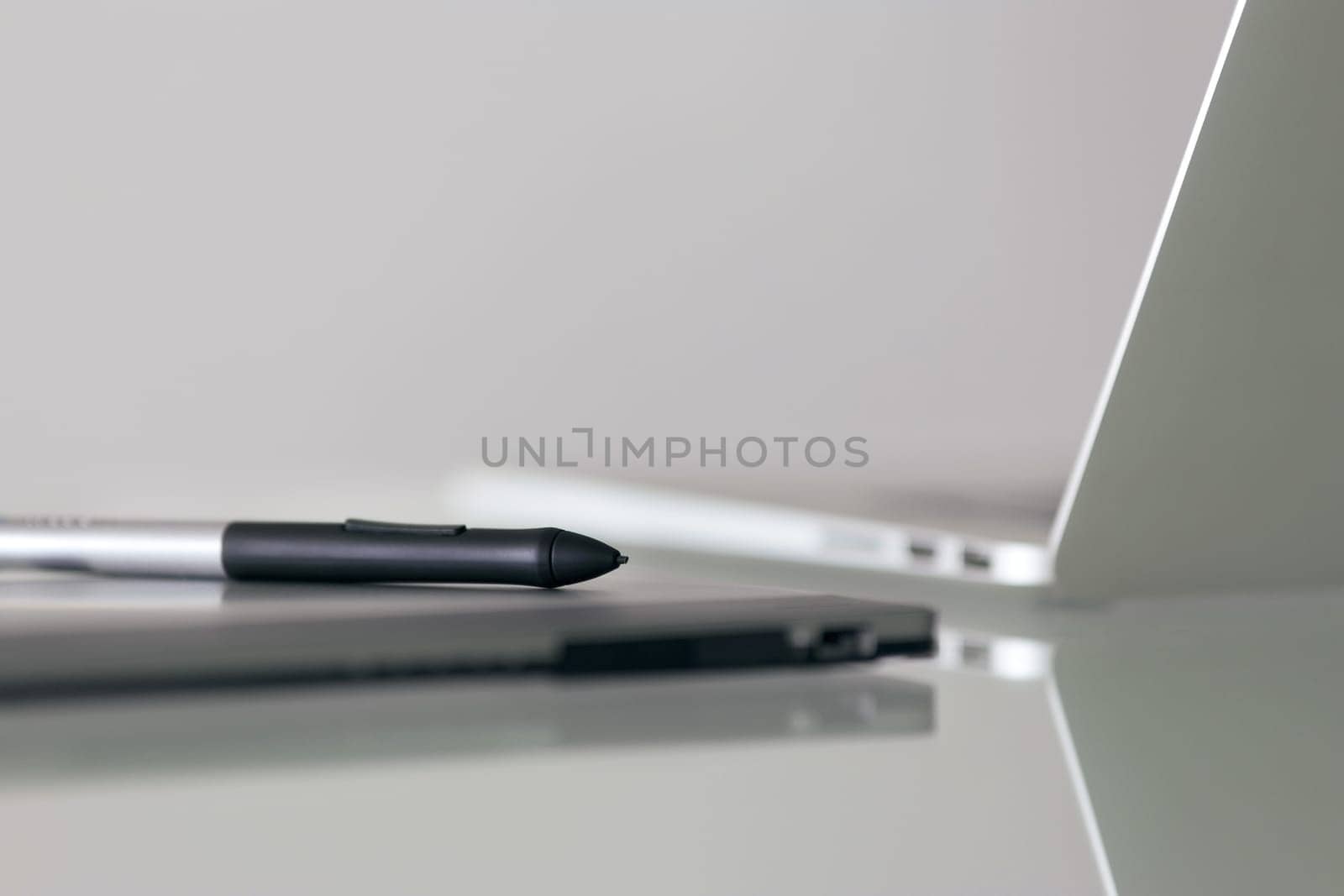 Still life of digital graphic tablet and stylus pen on desk, with laptop computer in background. Reflections on table and copy space on top gray wall