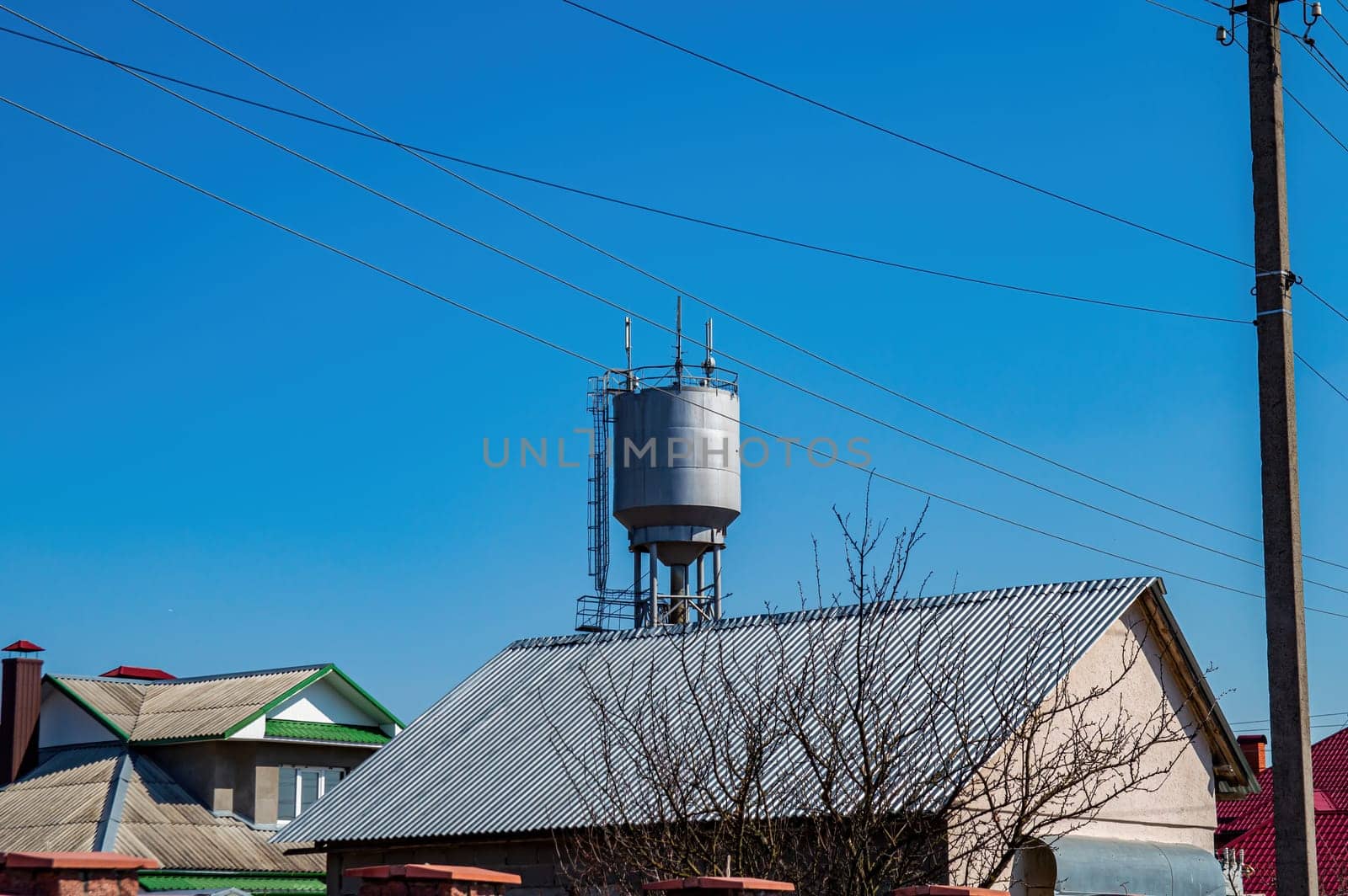 A water tower over the roofs of rural houses against the blue sky. Water tower. Roofs of houses. Blue sky. Water supply in the village. Metal industrial construction. Background image.