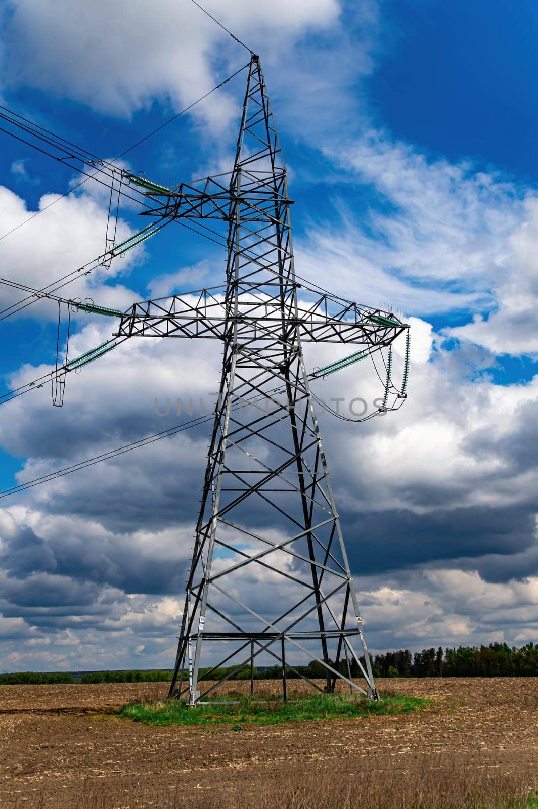 High-voltage power transmission tower with wires. High voltage line. Energy industry. Energy transfer. Blue sky. White clouds. Agricultural field. Cloudy horizon. Business. Electrician work.