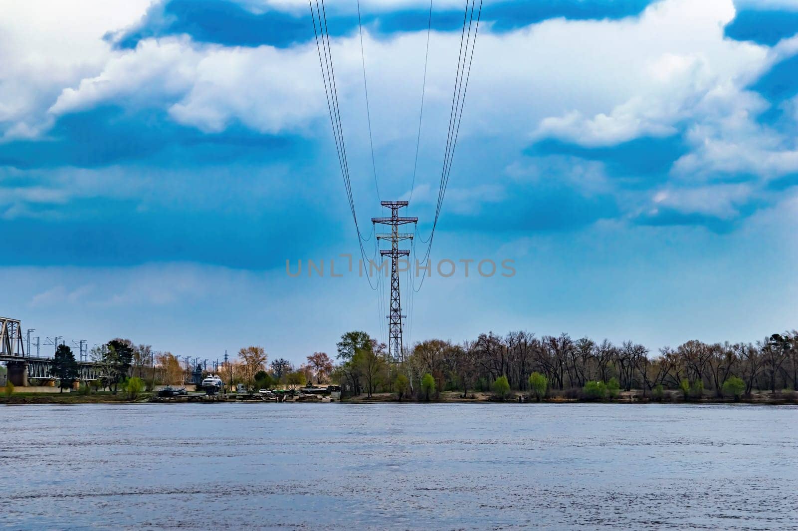 Wires of a high-voltage power line tower over a river. Electric wire. High voltage tower. Electric power transmission cable. Energy business. Dnieper river in Kyiv. Overcast sky. Industry.
