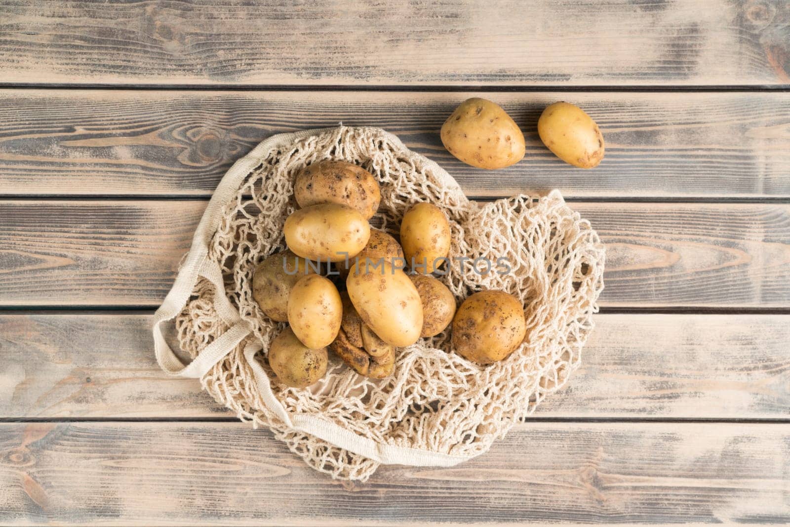 Bunch of unpeeled yellow new potatoes on faded wooden table, top view. Fresh farm vegetables in eco fabric mesh bag. Organic food or healthy diet concept. Planking. Culinary and cooking.