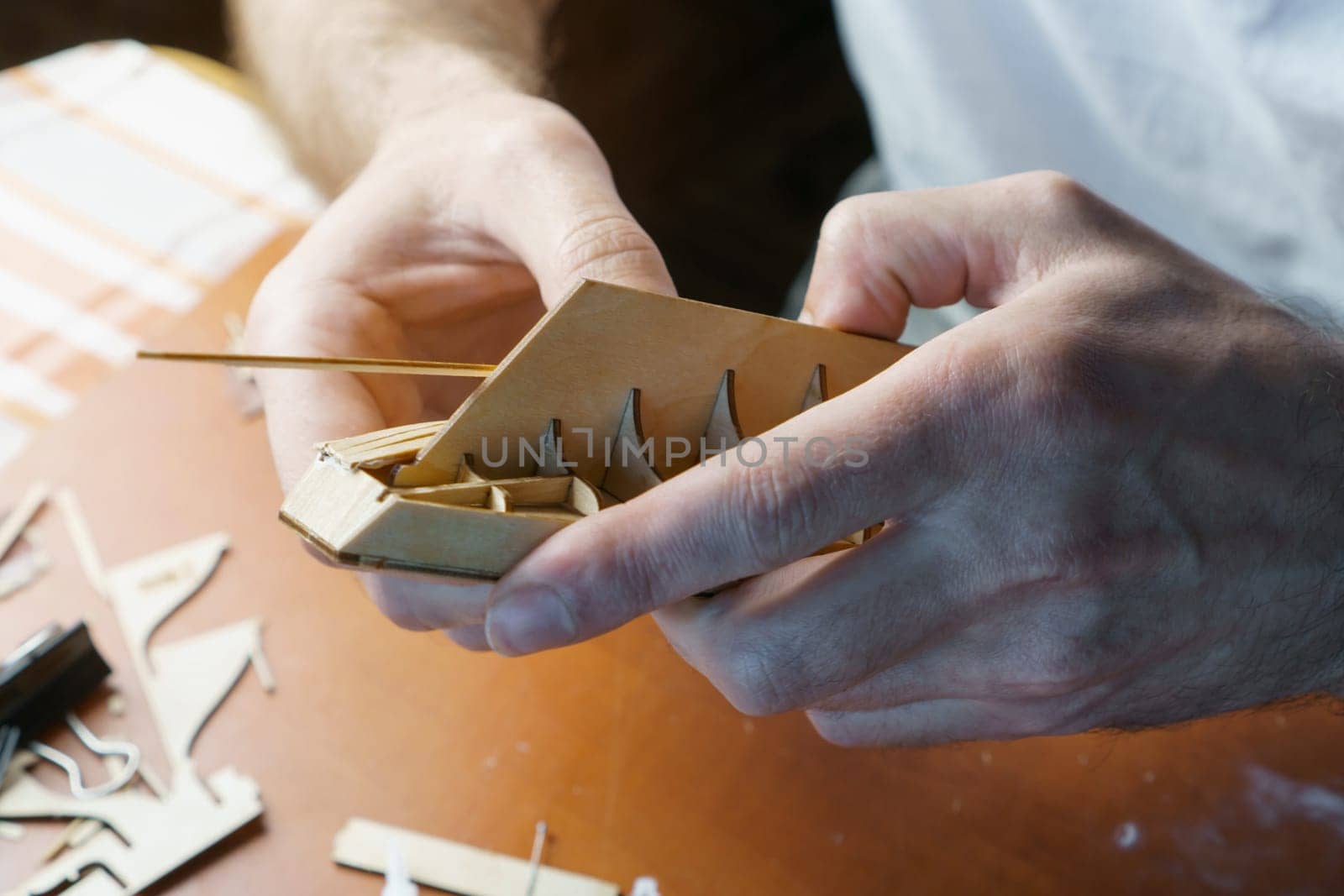 Hands of man gluing plywood details for ship model with glue, holding with fingers. Process of building toy ship, hobby, handicraft. Table with various materials, parts and devices for work