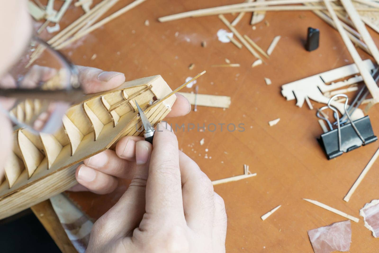 Hands of man cutting out details for ship model with clerical knife from plywood. Process of building toy ship, hobby and handicraft. Table with various materials, parts and devices for work