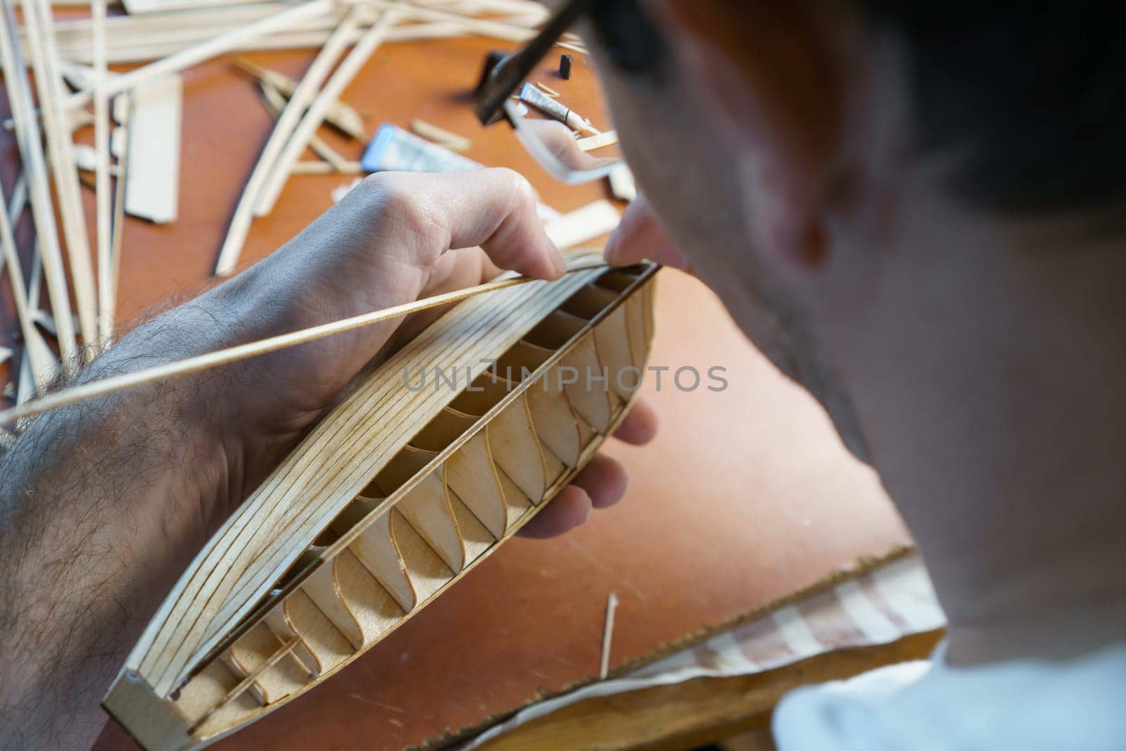 Hands of man gluing plywood details for ship model with glue, holding with fingers. Process of building toy ship, hobby, handicraft. Table with various materials, parts and devices for work