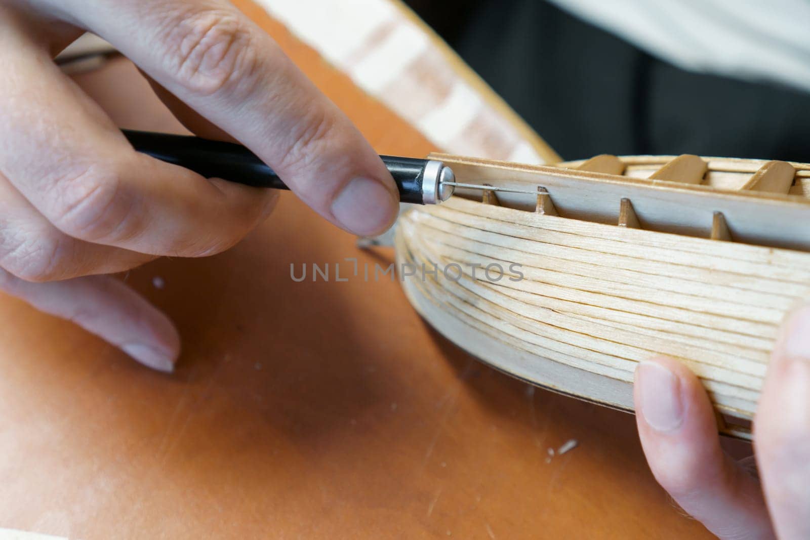 Hands of man removing excess glue from plywood details for ship model with utility knife. Process of building toy ship, hobby, handicraft. Table with various materials