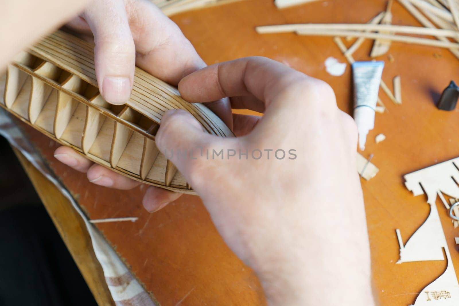 Hands of man gluing plywood details for ship model with glue, holding with fingers. Process of building toy ship, hobby, handicraft. Table with various materials, parts and devices for work