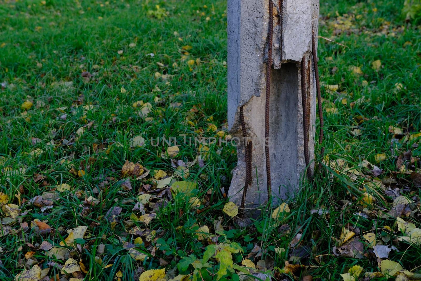 A close-up view of a dilapidated concrete column battered by an accident on the side of a road with green leaves and weeds.
