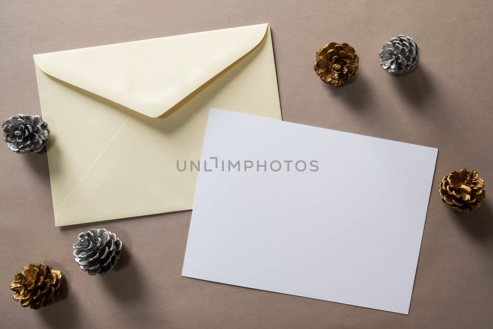 Christmas winter festive ornament. Blank paper card and envelope with silver and golden pine cones on brown background. Minimal decoration. Invitation Christmas holiday party. Flat lay, top view, copy space.