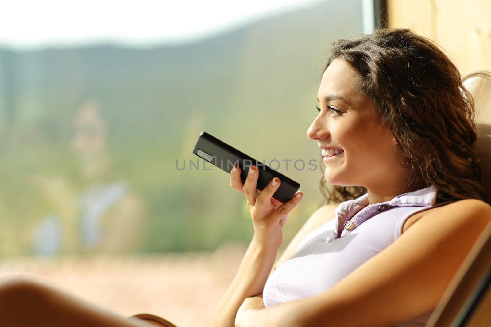 Happy woman sitting on a chair dictating message on smart phone looking through a window at home