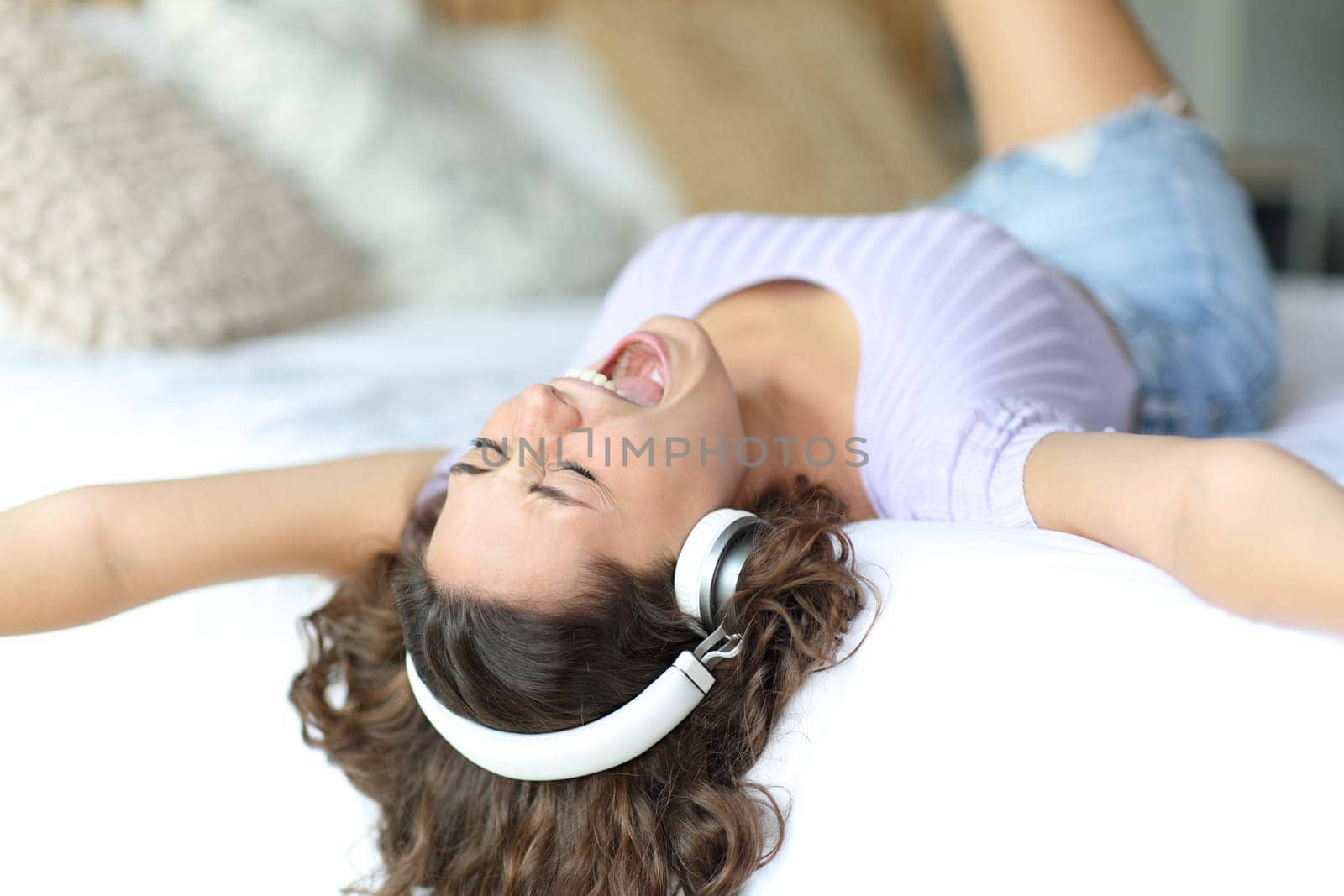 Excited woman singing and listening to music wearing headphones lying on a bed in a room