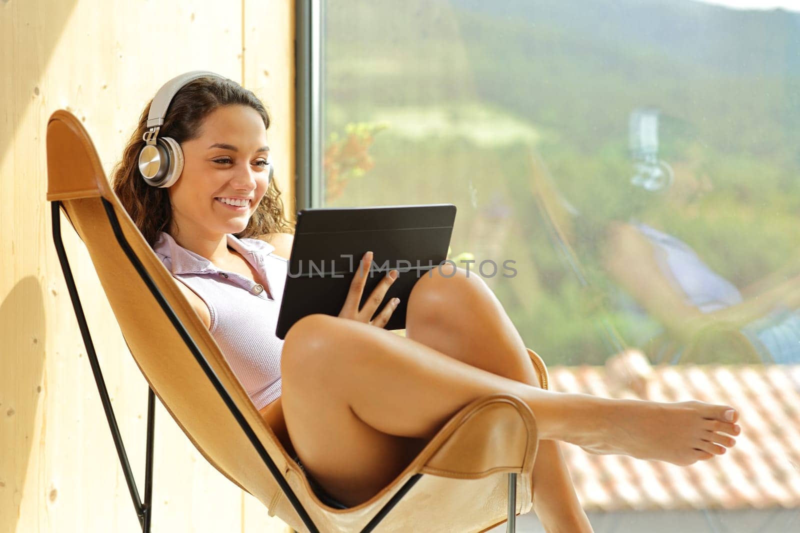 Happy woman watching media with tablet and headphones sitting on a chair at home