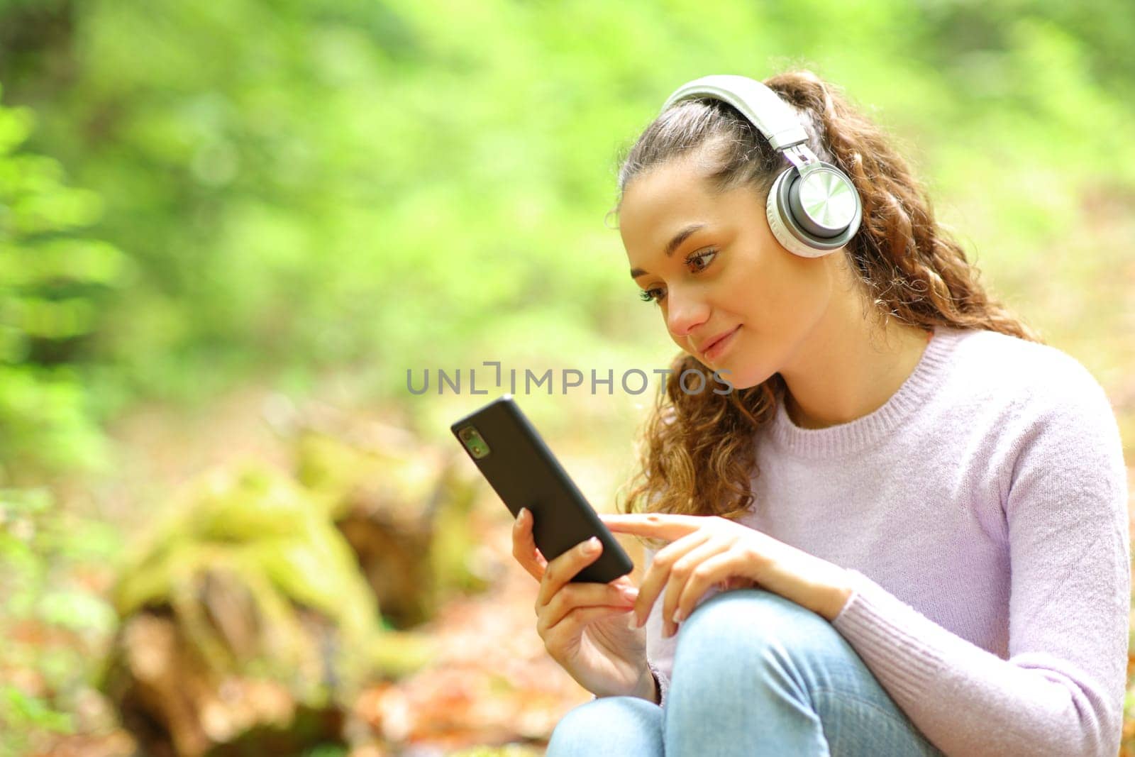 Relaxed woman wearing headphones listening music and wathing phone content in a forest