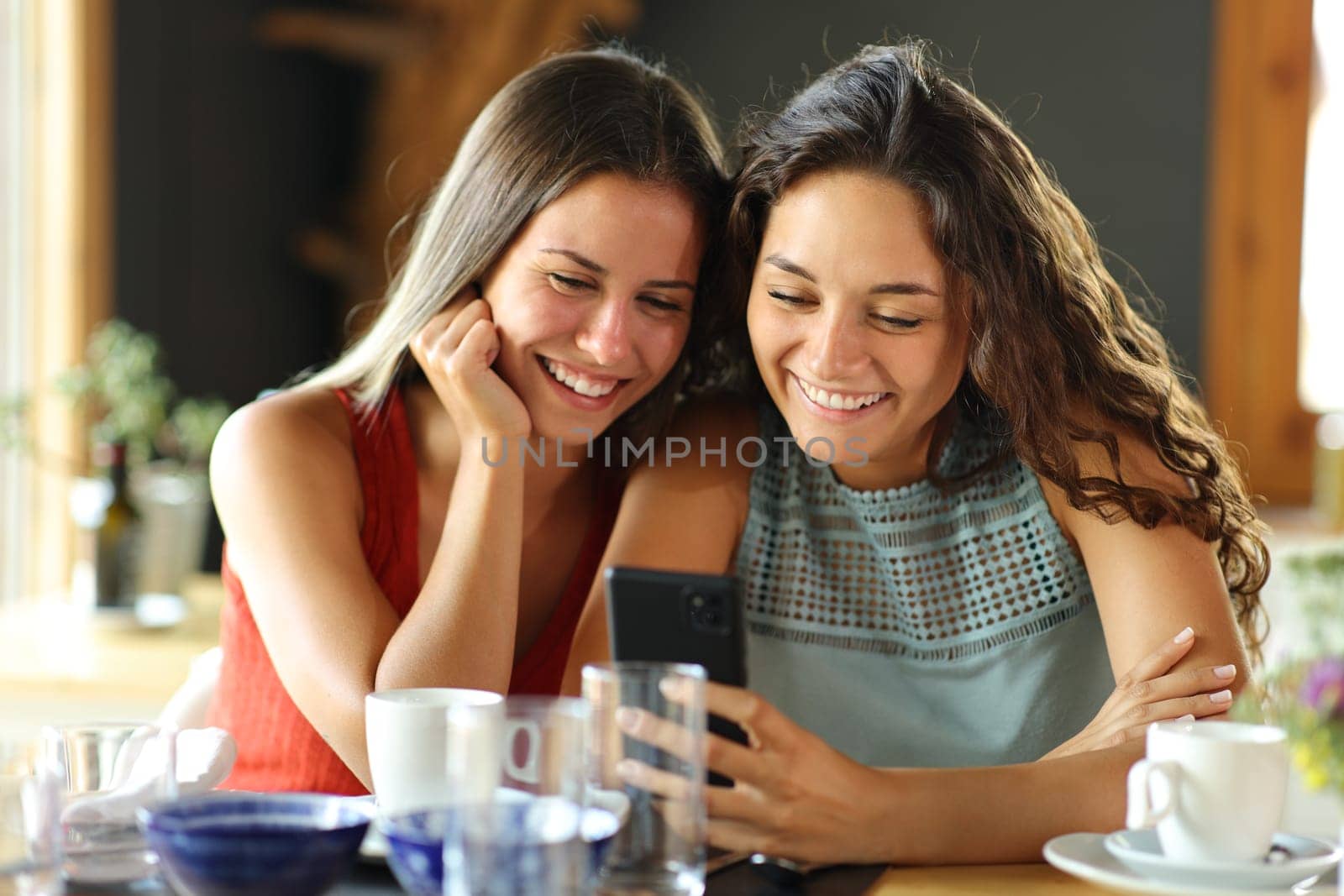 Front view of two happy friends checking smart pone in a restaurant