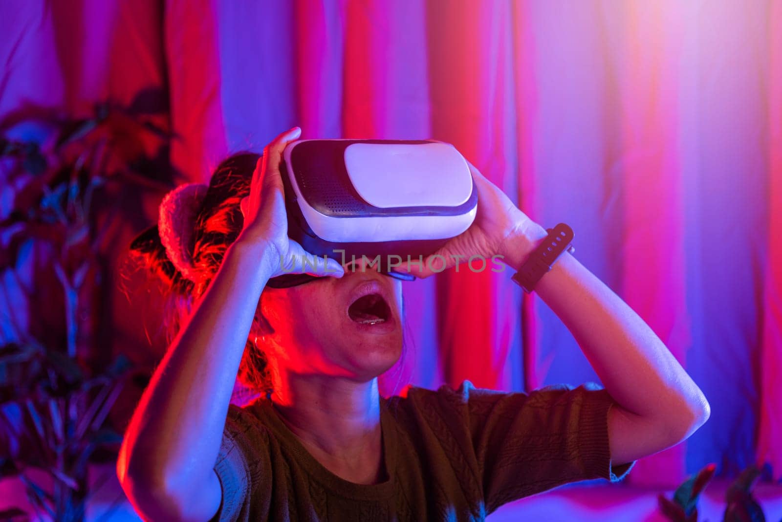Asian woman wear the VR helmet excited emotional at home purple and blue background, female experiencing virtual reality goggles experiencing reality, metaverse and cyberspace concept