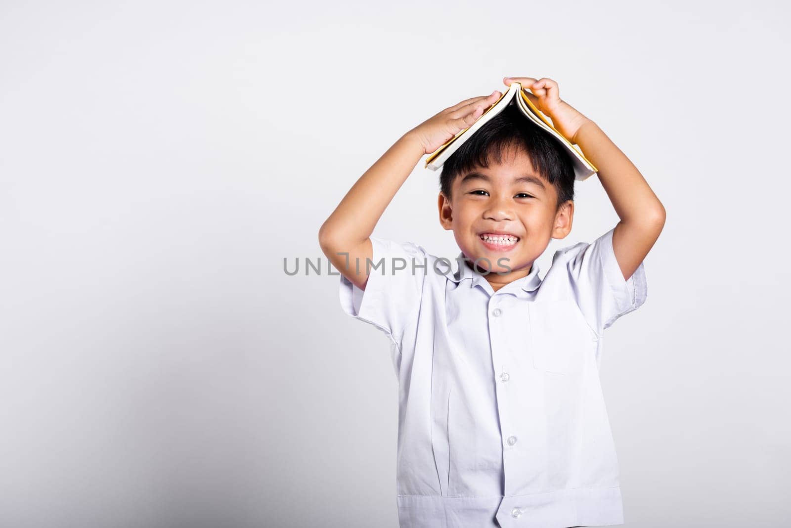 Asian adorable toddler smiling happy wearing student thai uniform red pants stand holding book over head like roof in studio shot isolated on white background, Portrait little children boy preschool