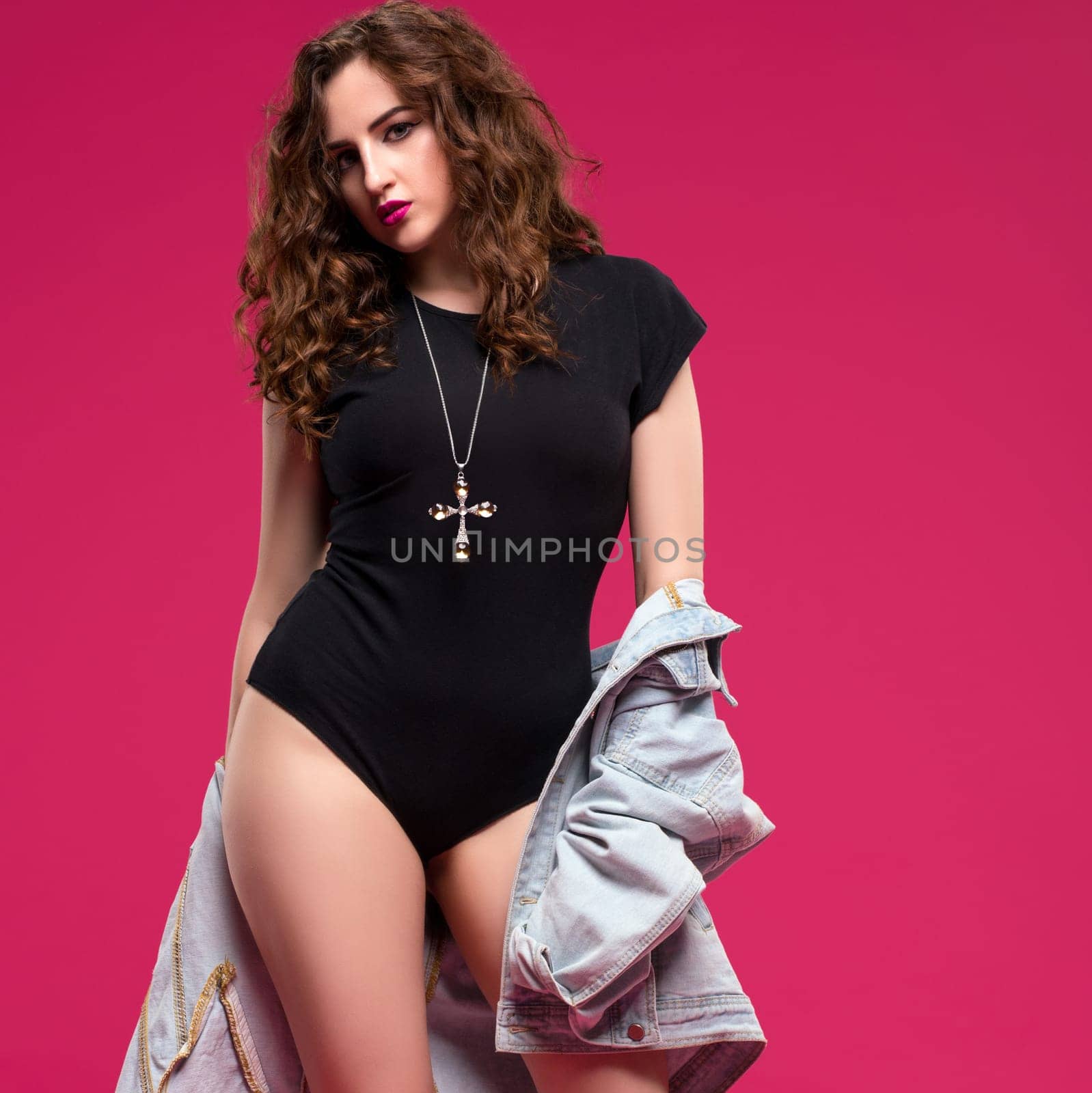 A young model showcases a stylish black bodysuit with a denim jacket, exuding confidence in front of a bright pink background.