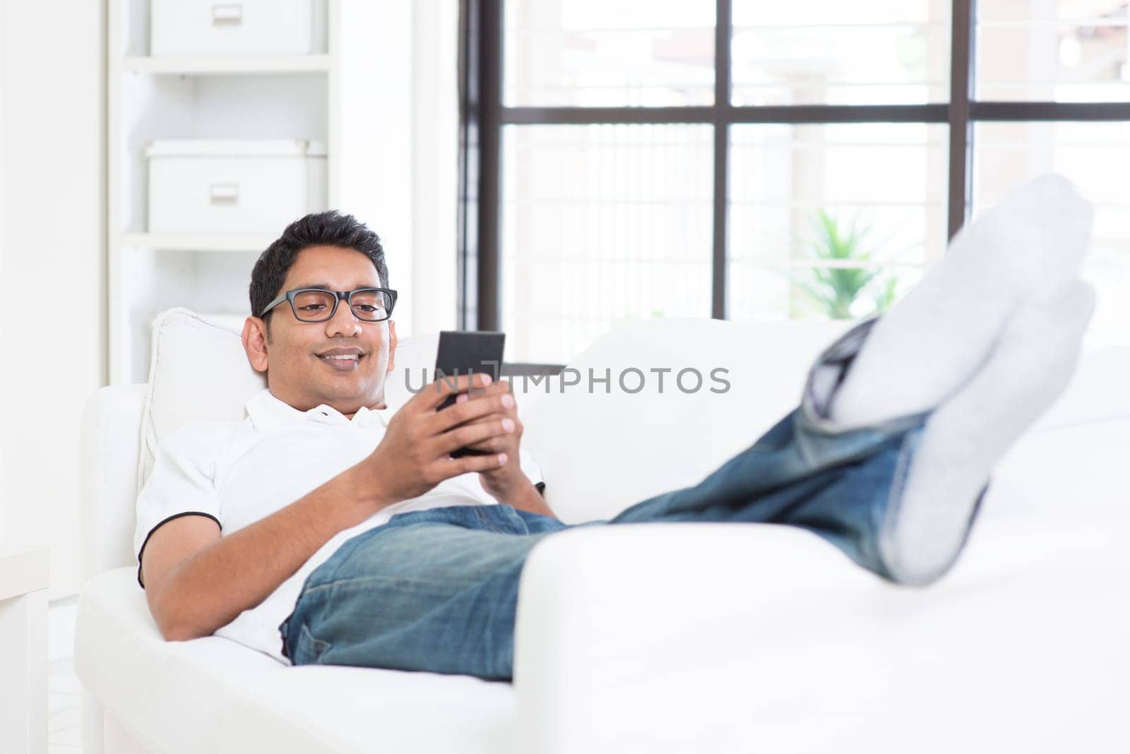 Smartphone mobile apps concept. Indian guy using smart phone. Asian man relaxed and lying on sofa indoor. Handsome male model.
