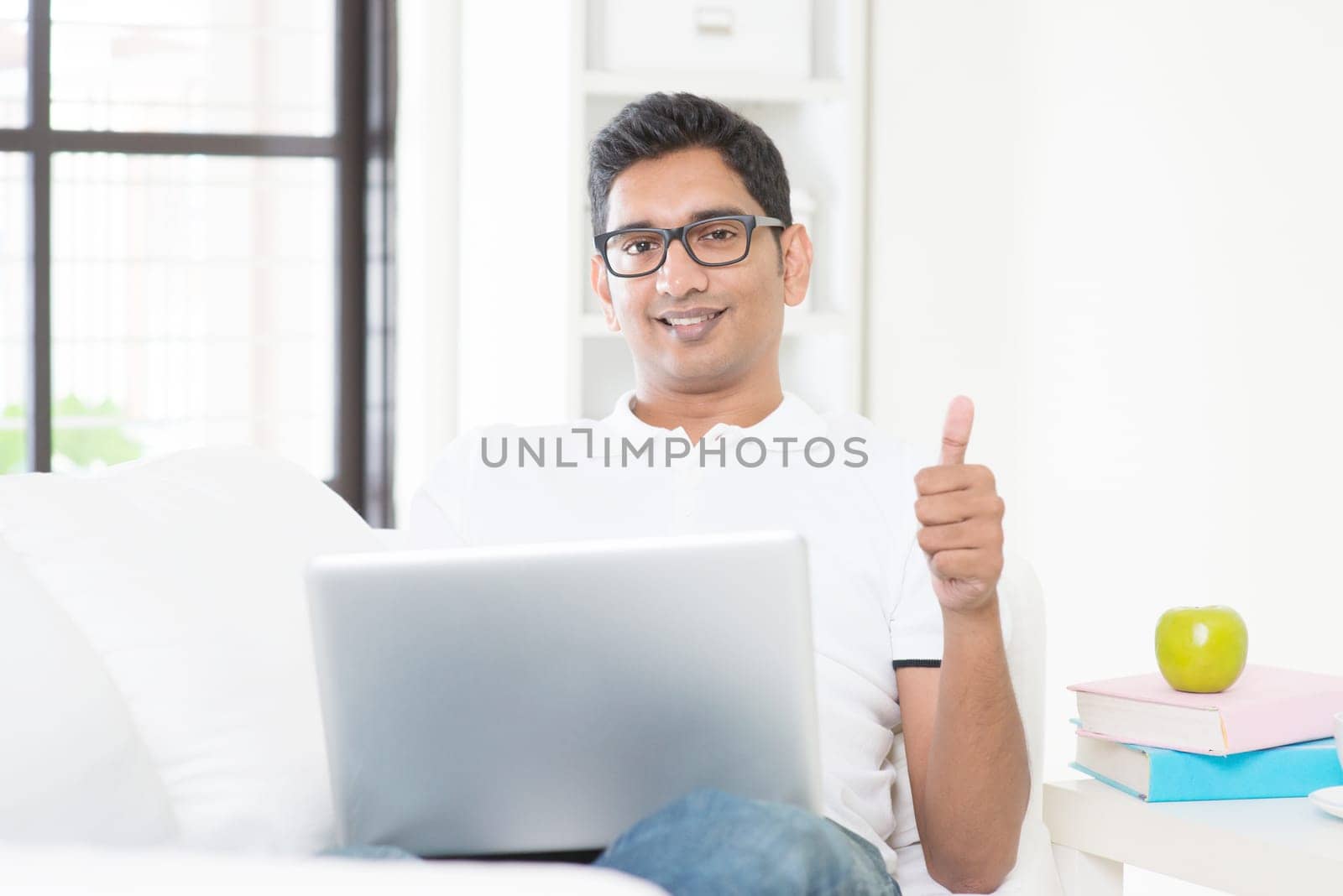 Indian guy with laptop computer working from home and giving thumb up. Asian man using internet indoor, relaxed and sitting on sofa. Handsome male model.
