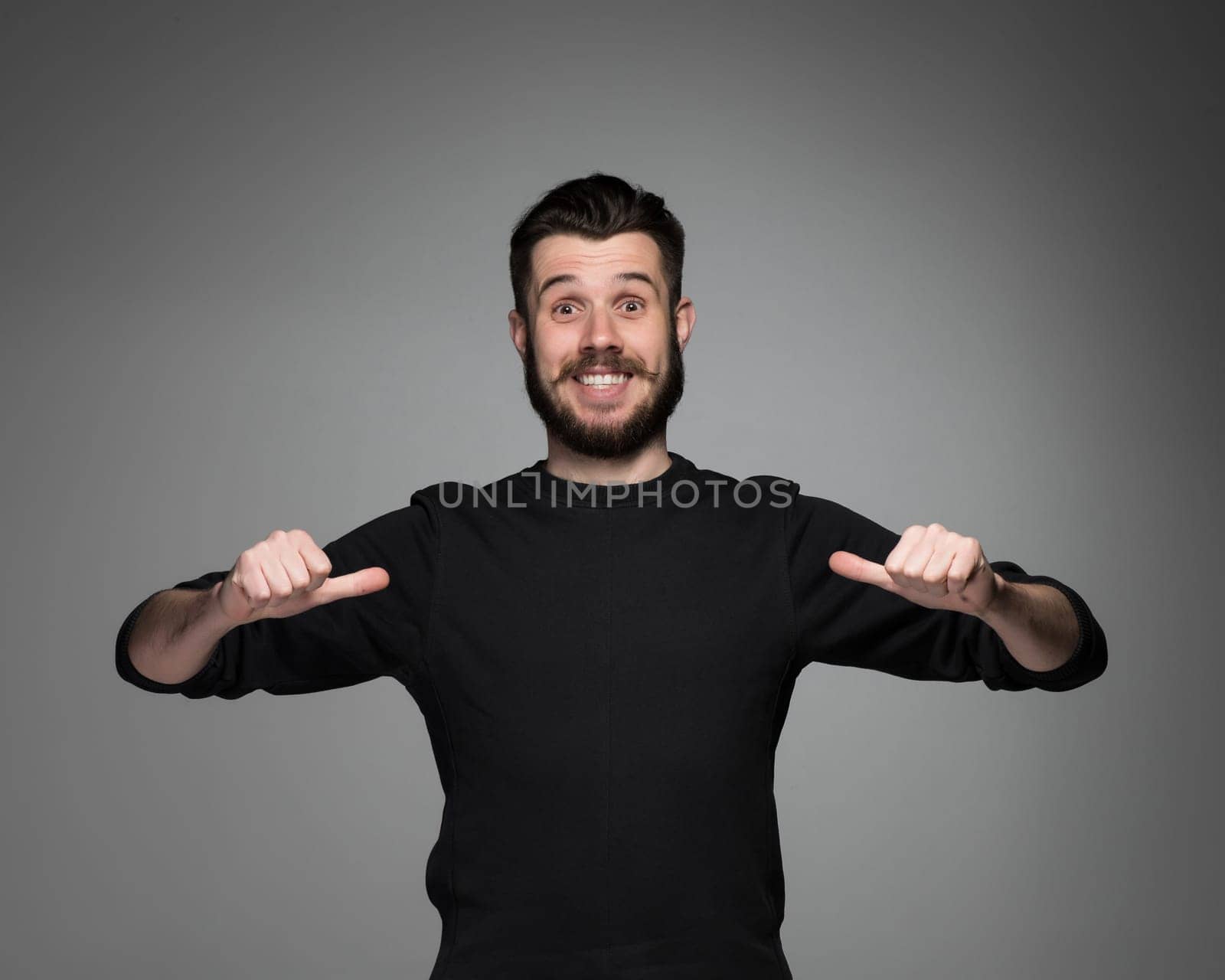 young smiling man with a beard and mustache in black pointing himself on a gray background