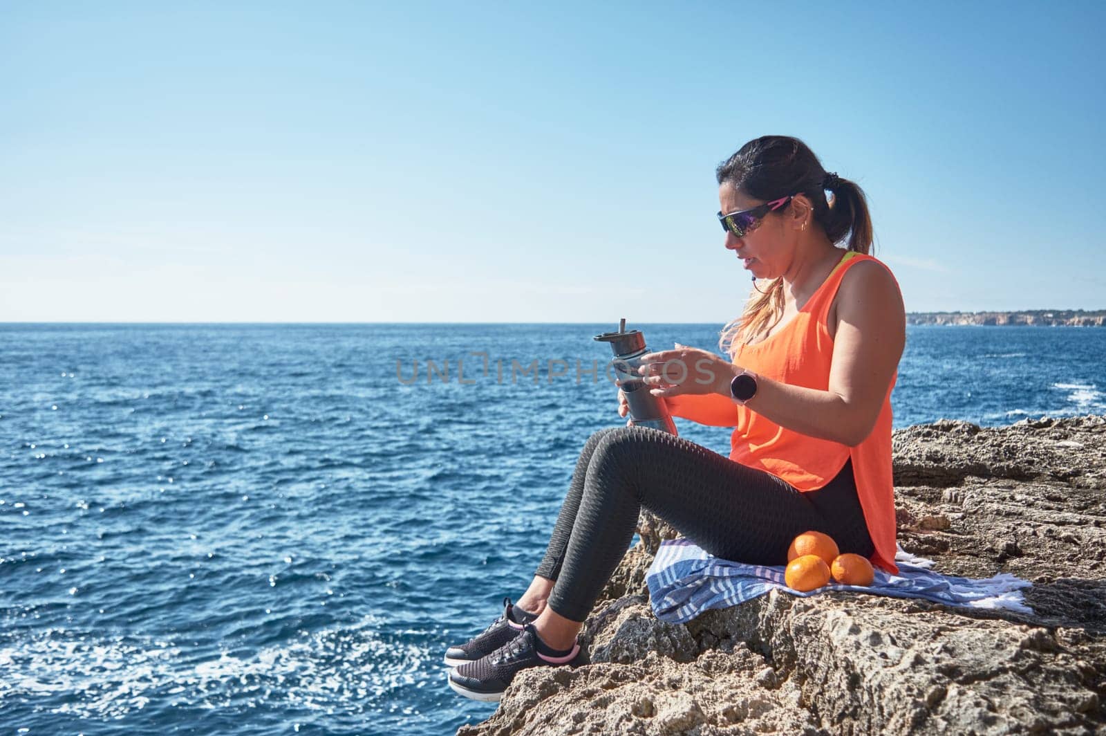 Latin woman, middle-aged, resting, regaining strength, eating, drinking water, after a gym session, burning calories, keeping fit, outdoors by the sea, wearing headphones and smart watch