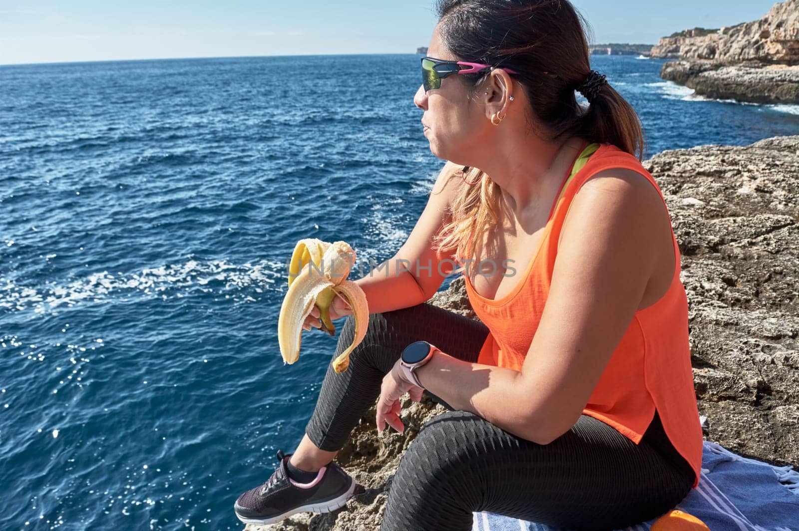Latin woman, middle-aged, resting, regaining strength, eating, drinking water, after a gym session, burning calories, keeping fit, outdoors by the sea, wearing headphones and smart watch