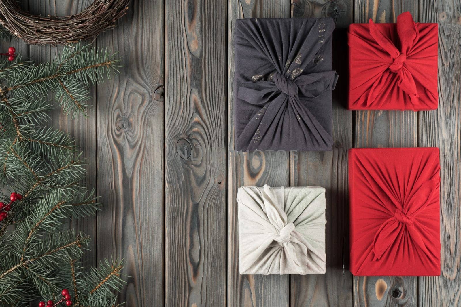 Holiday composition four handmade, craft, wrapped Furoshiki present boxes with decorated fir tree branches and wreath on dark wooden background. Holiday tradition for Christmas. Flat lay, copy space