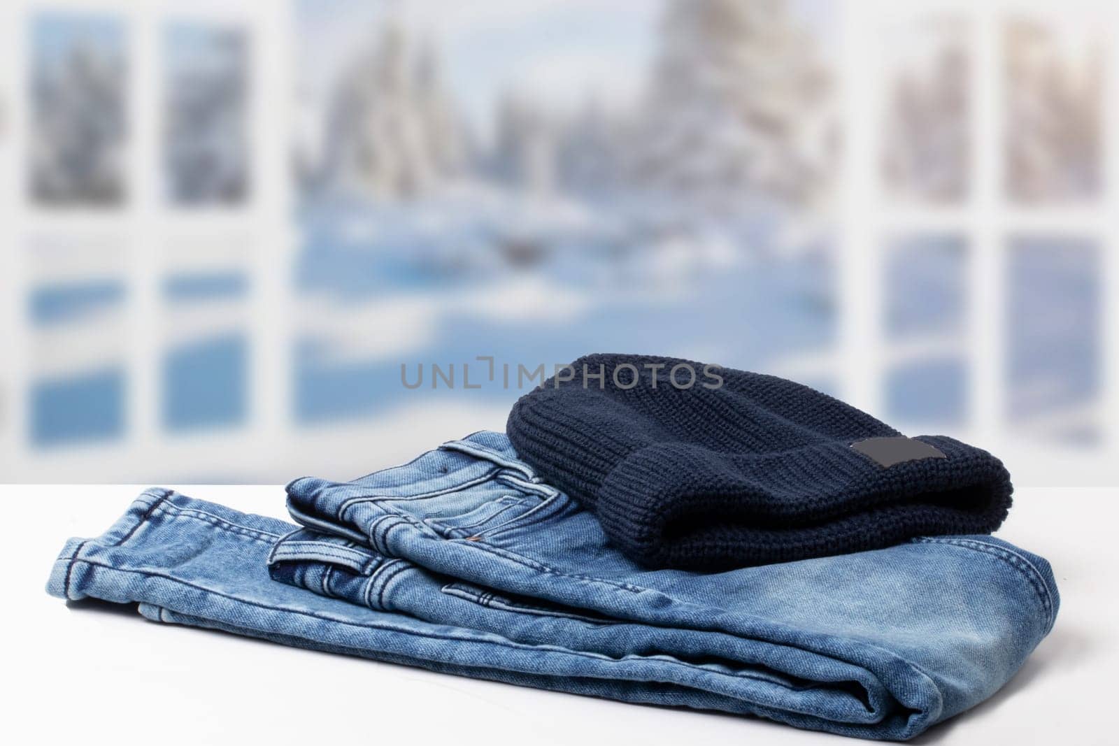 Autumn clothes. Fashionable folded jeans and a knitted blue Rapper, Beanie or Baseball hat or cap on table over abstract blurred winter landscape. Copy space.
