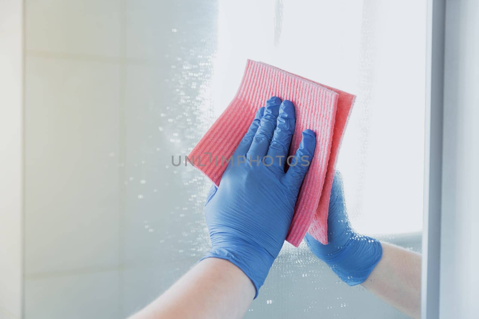 Cropped view of housekeeper woman hand in blue rubber glove polishing, cleaning, swiping mirror with cloth rag, spraying cleaner liquid while cleaning bathroom at home. Housekeeping, cleaning service