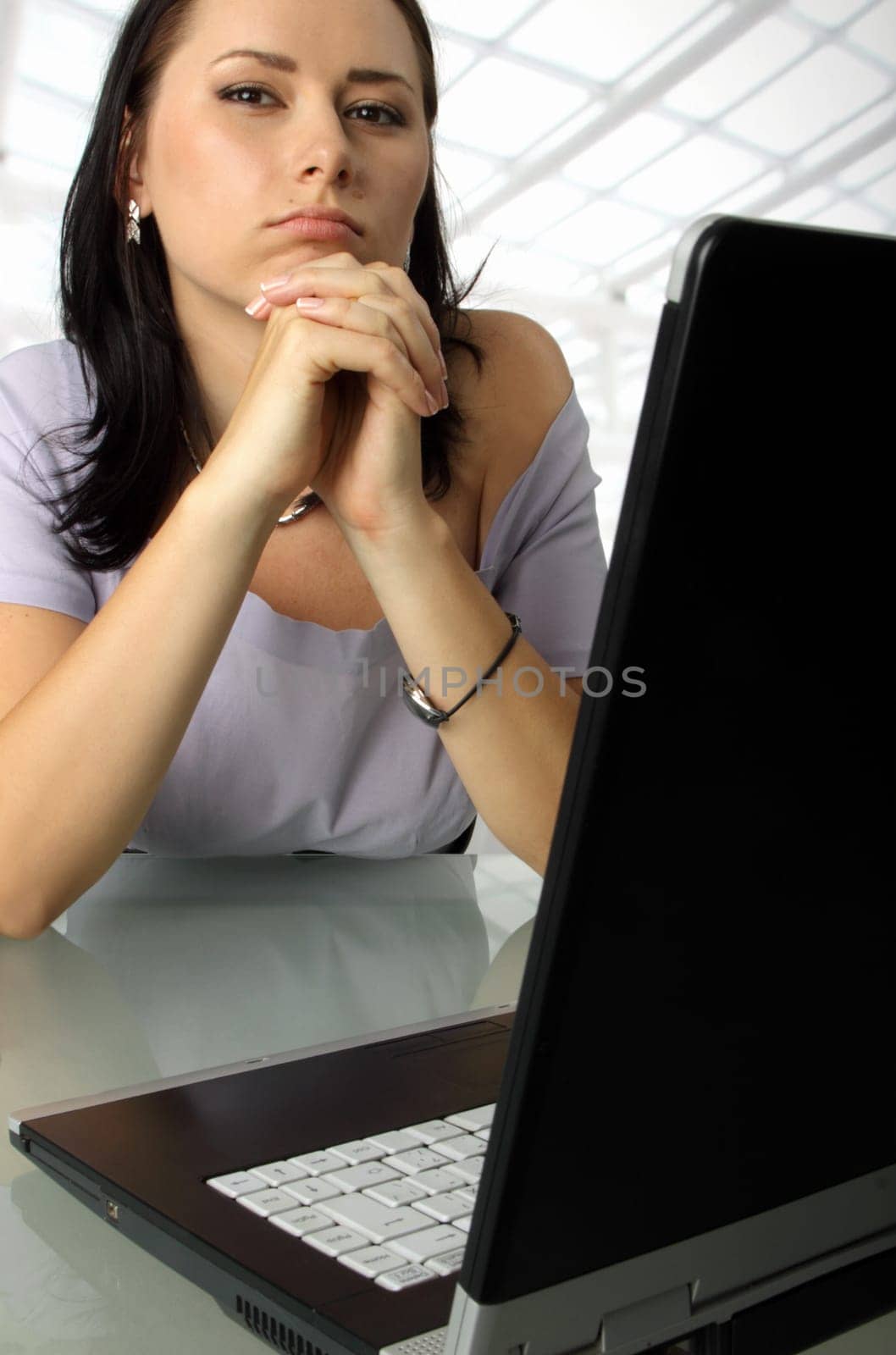 Serious female is sitting behind a laptop thinking, fingers crossed.
