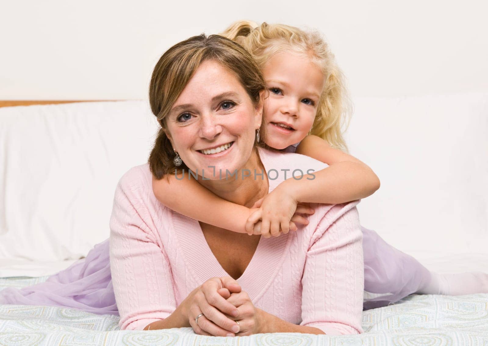A mother is lying with her daughter on her bed.  The girl has her arms aroung her mother and they are smiling at the camera.  Horizontally framed shot.