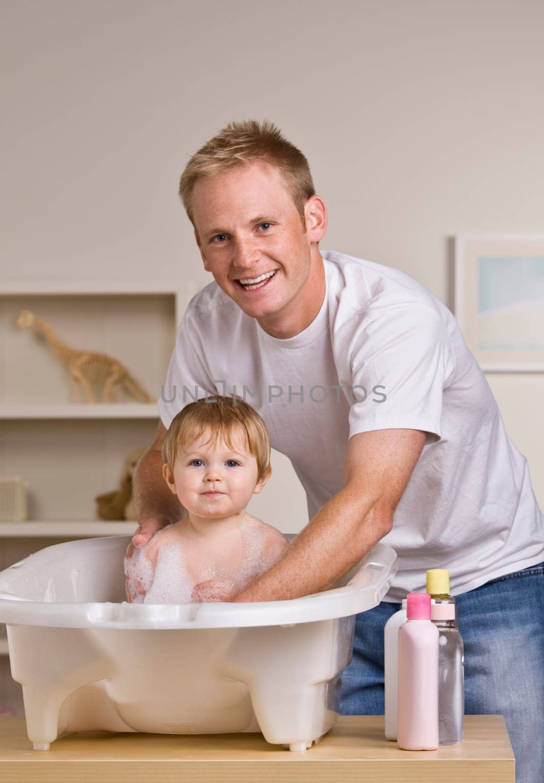 A young father is giving his baby daughter a bath in a childs bathtub.  He is smiling at the camera.  Vertically framed shot.