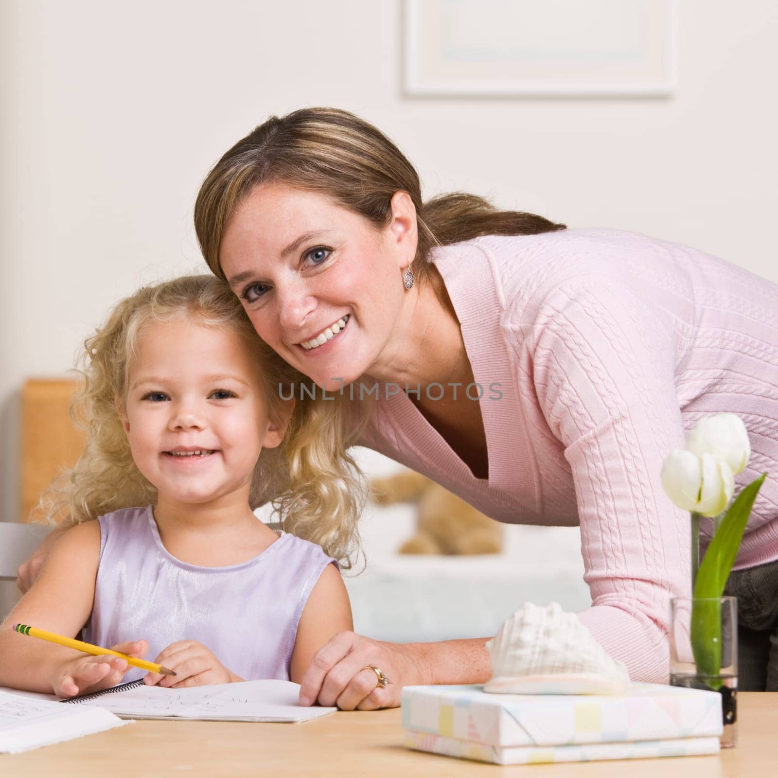 A mother is sitting with her young daughter and watching her draw.  They are smiling at the camera.  Square framed shot.