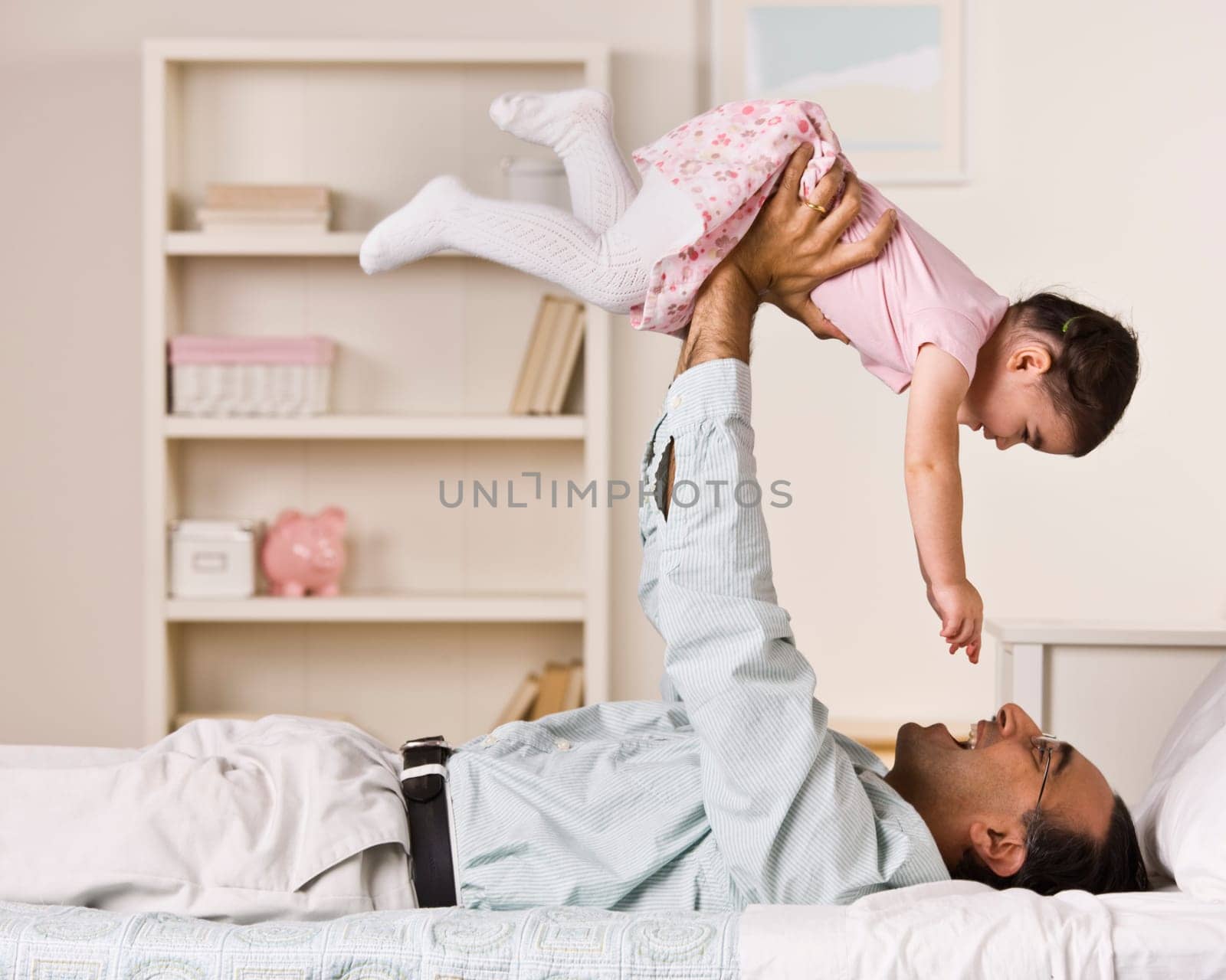 A father is holding his young daughter in the air above him.  They are smiling at each other.  Square framed shot.