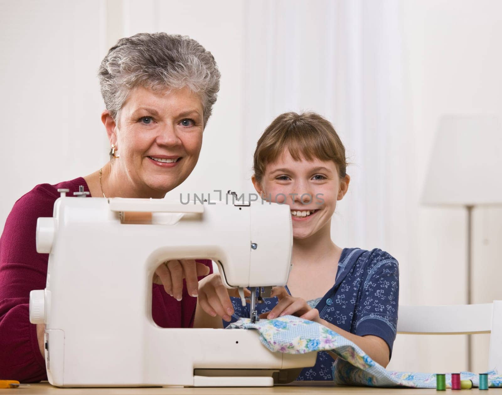 A young girl is sewing with her grandmother in the living room.  They are smiling at the camera.  Square framed shot.