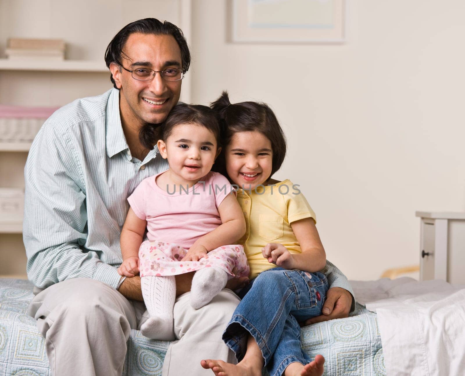 A father is sitting on a bed with his two young daughters.  They are smiling at the camera.  Horizontally framed shot.