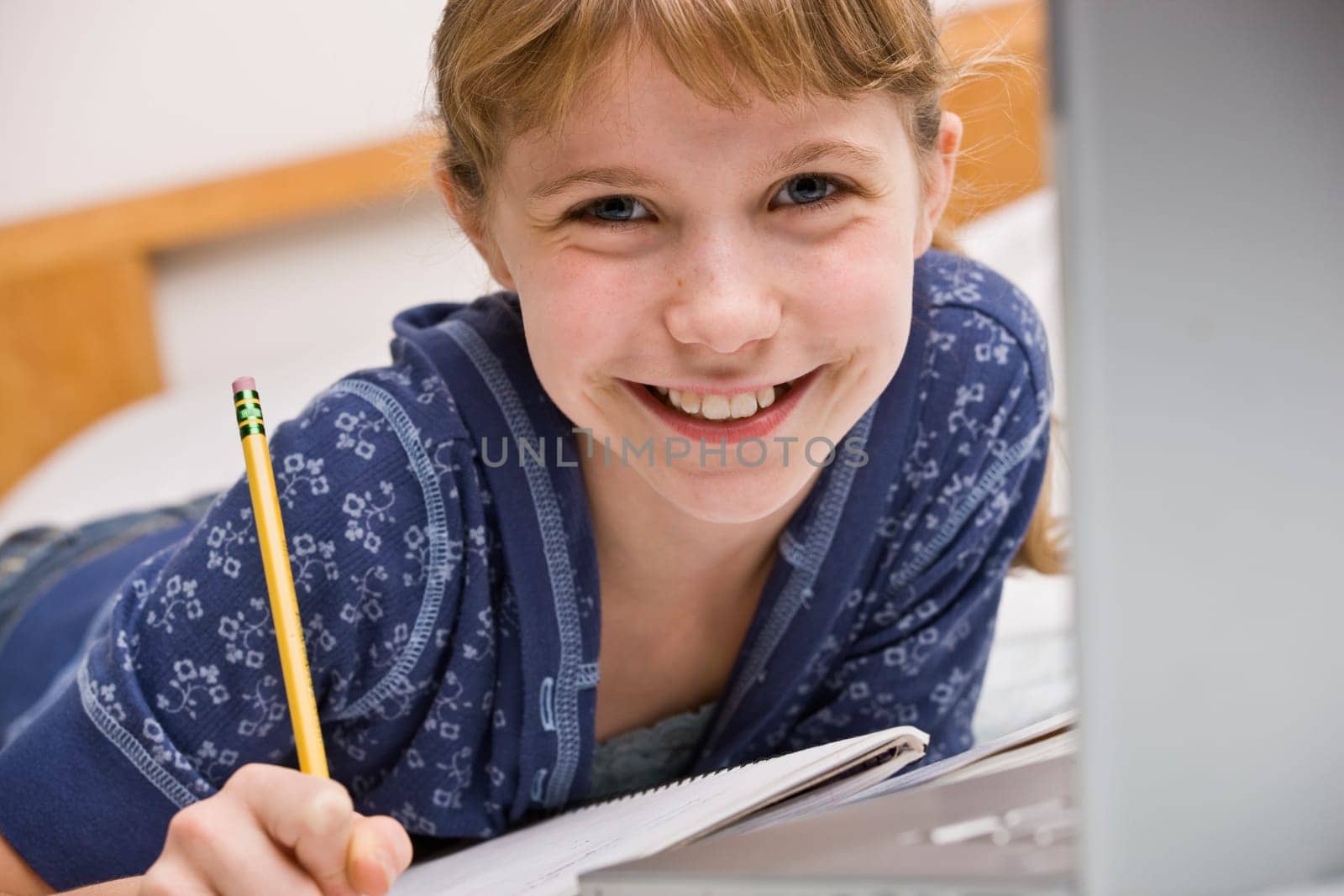 A young girl is seated at a computer desk and is working on her homework.  She is smilng at the camera.  Horizontally framed shot.