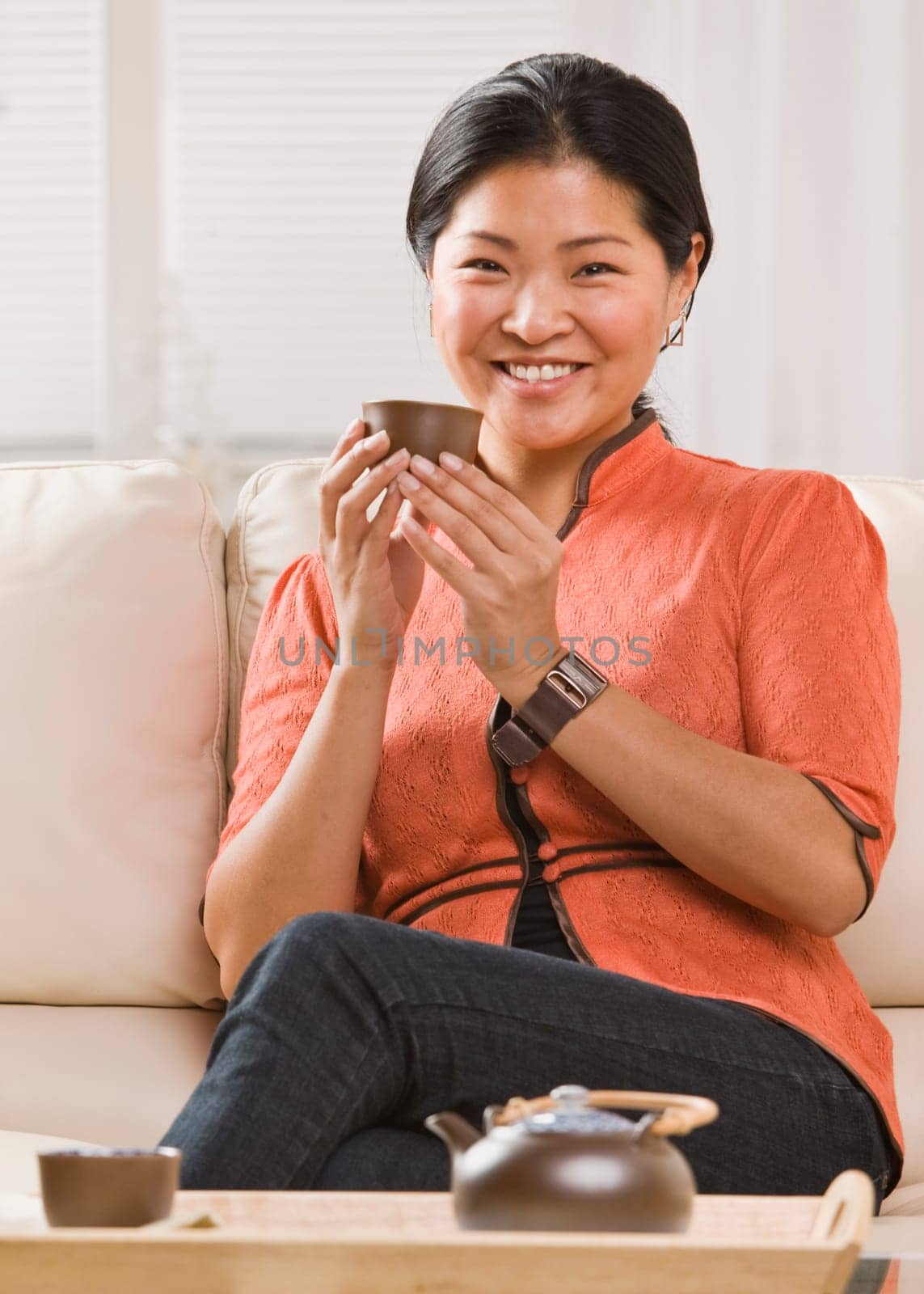 A woman is seated on her sofa, and drinking tea out of a tea cup.  She is smiling at the camera.  Vertically framed shot.