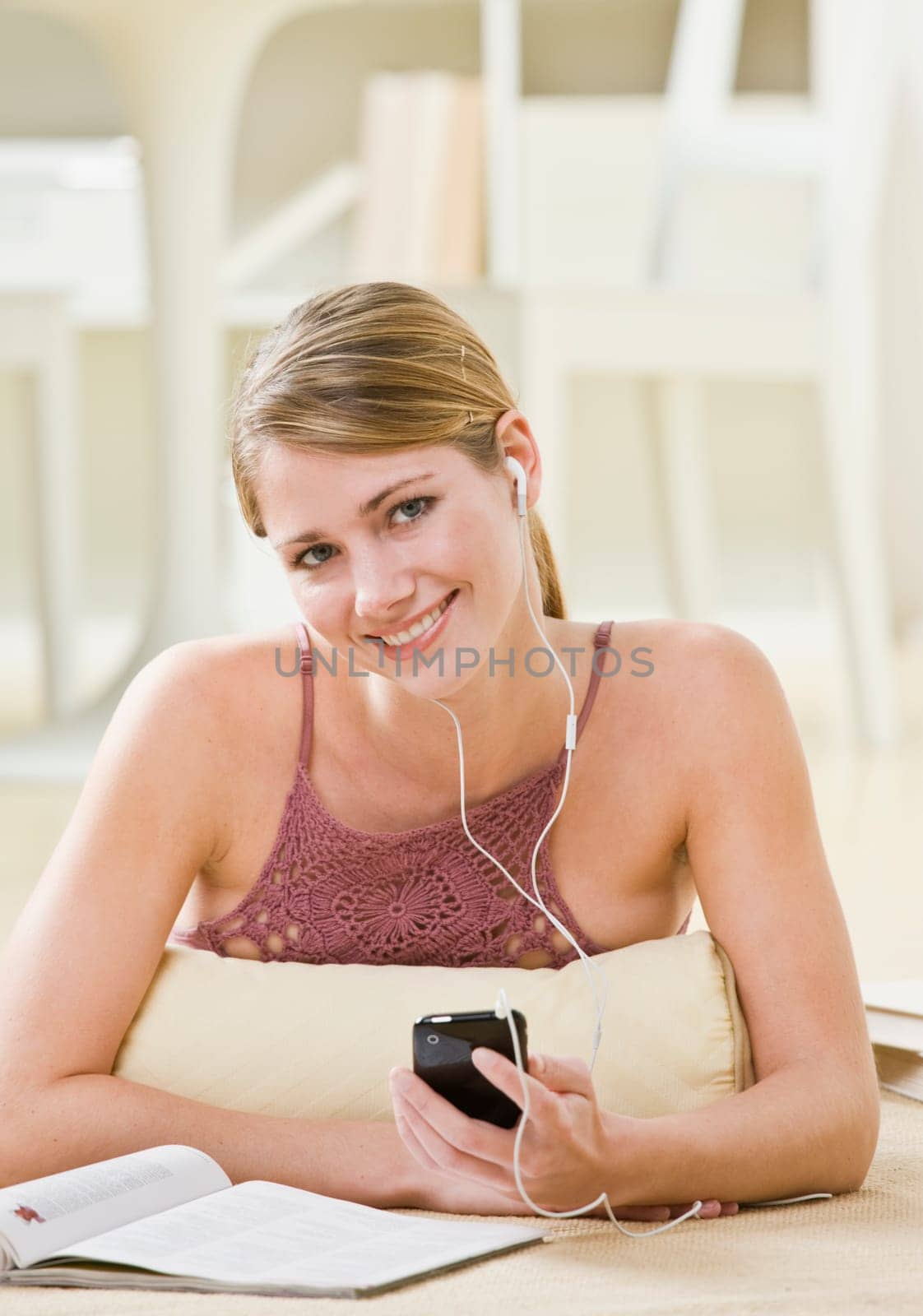 A young woman is laying down and listening to an MP3 player.  She is smiling at the camera.  Vertically framed shot.