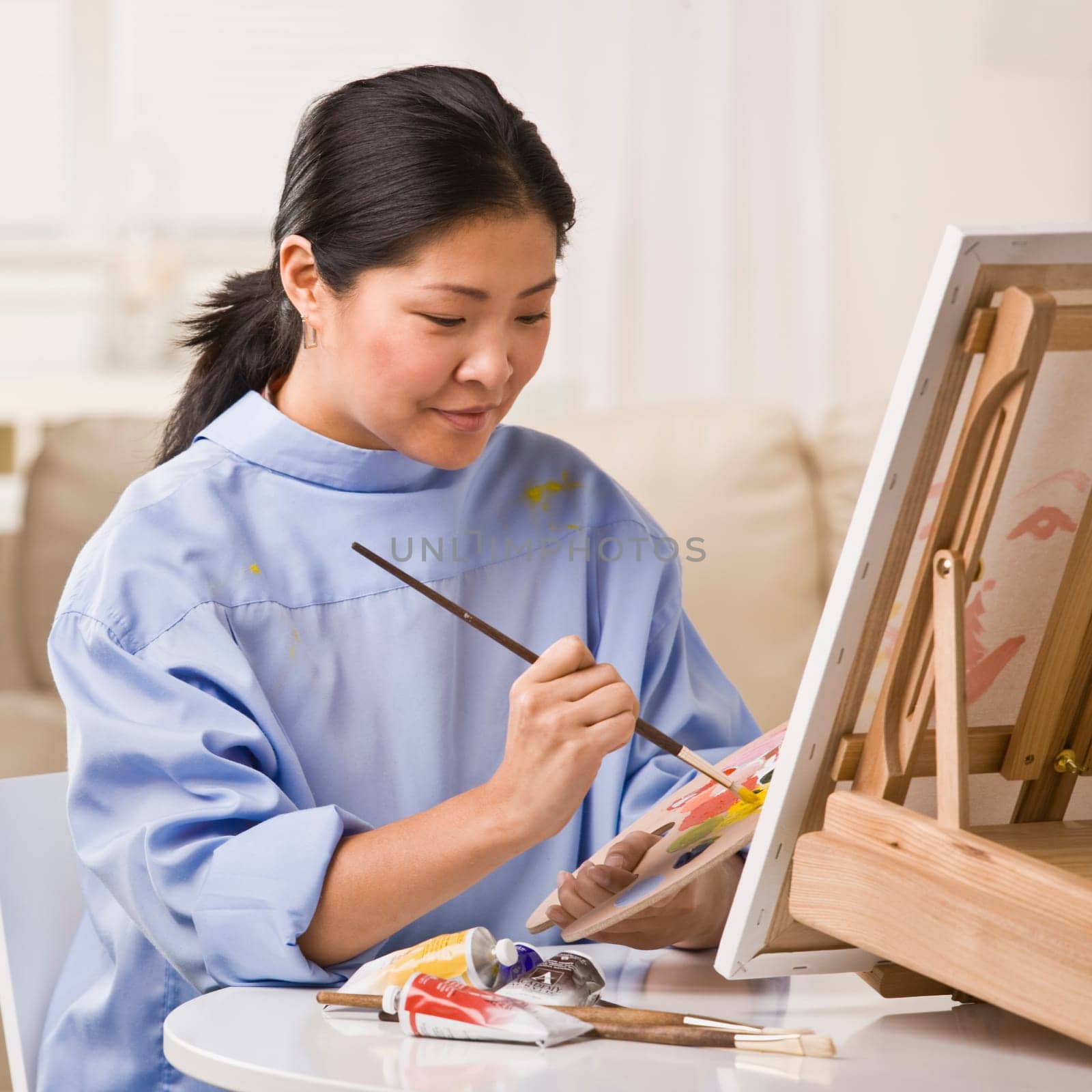 A woman is painting a picture on an easel.  She is looking away from the camera.  Square framed shot.