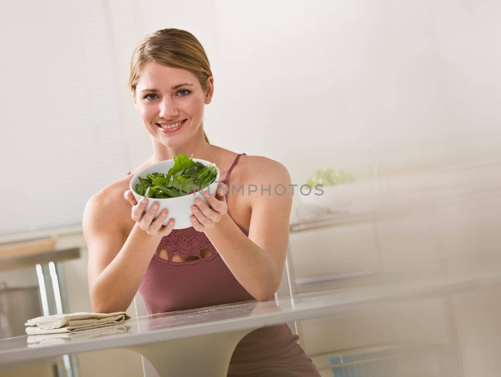 A young woman is seated in the kitchen and is holding out a bowl of salad.  She is smiling at the camera.  Horizontally framed shot.