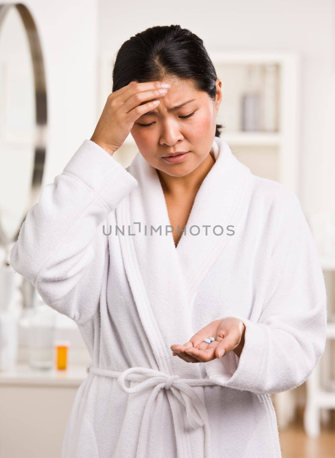 A young woman is holding pain medication that she is about to take for a headache.  She is looking away from the camera.  Vertically framed shot.
