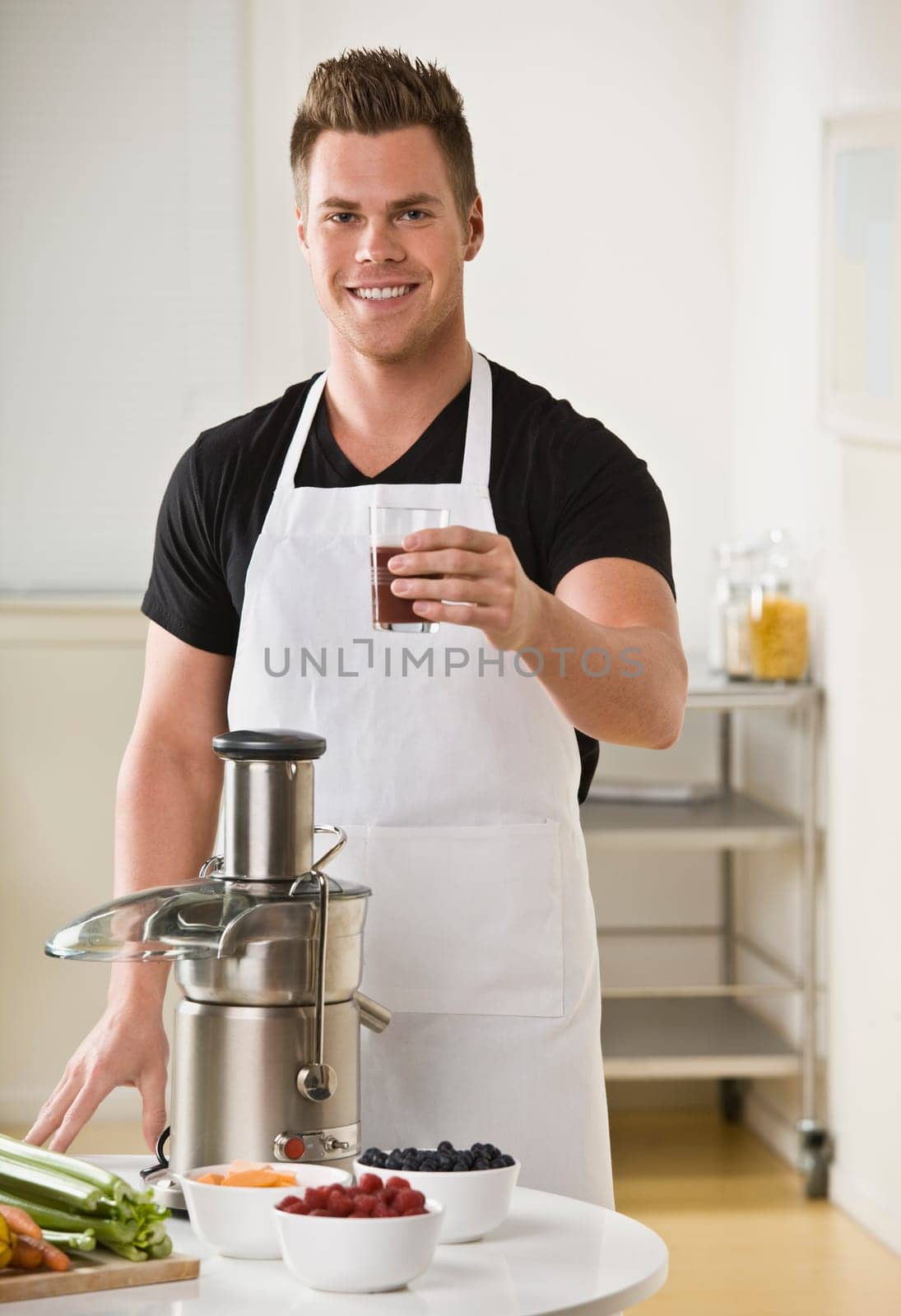 A young man is standing in his kitchen and making juice from a juicer.  He is smiling at the camera.  Vertically framed shot.