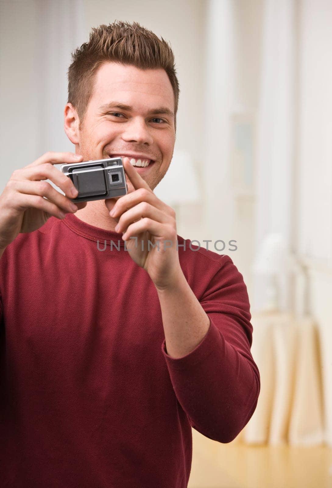 A young man is holding up a digital camera and smiling at the camera. Vertically framed shot.