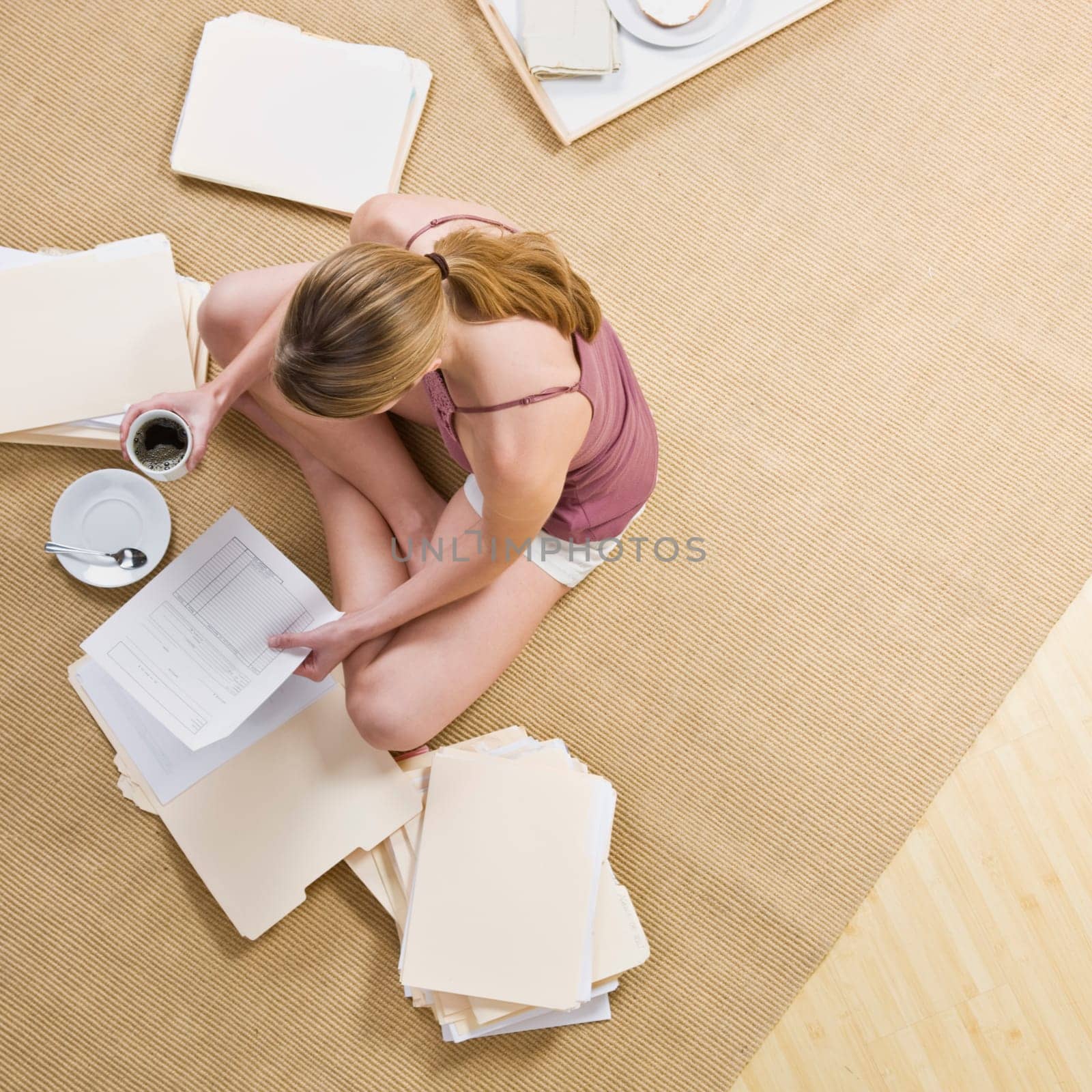 A woman is seated on the floor, looking through files, and drinking coffee.  Square framed shot.