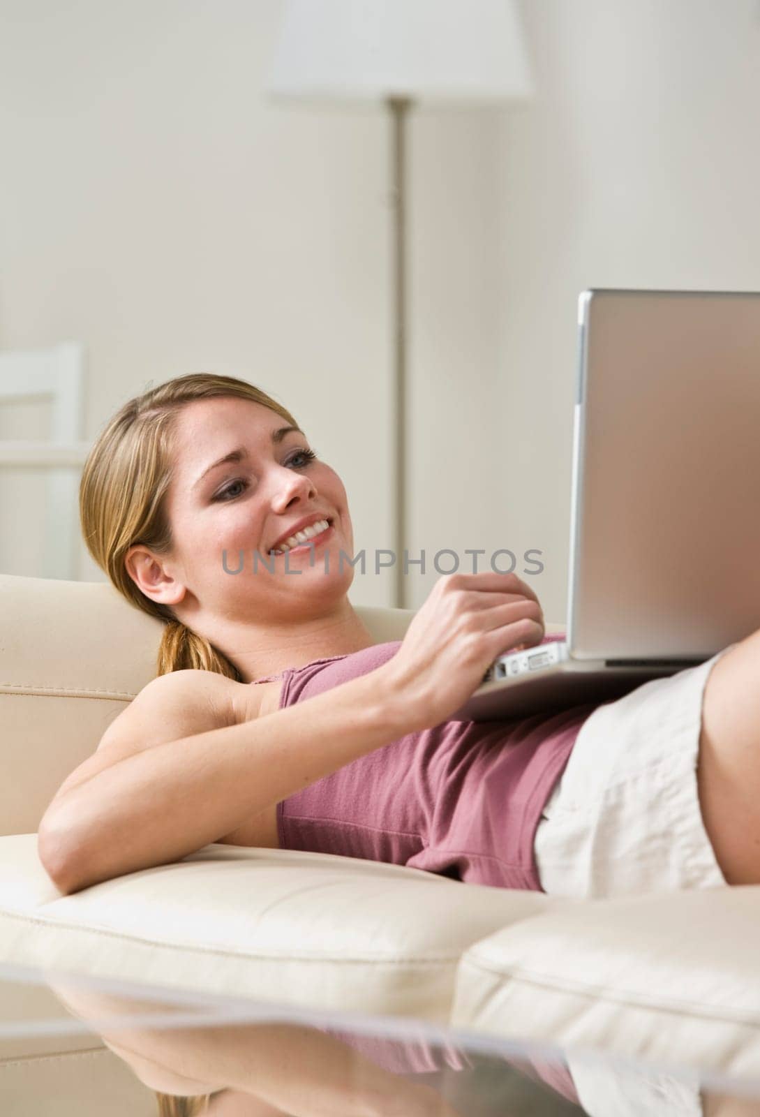 A young, smiling woman is laying on a couch and looking at a laptop.  Vertically framed shot.