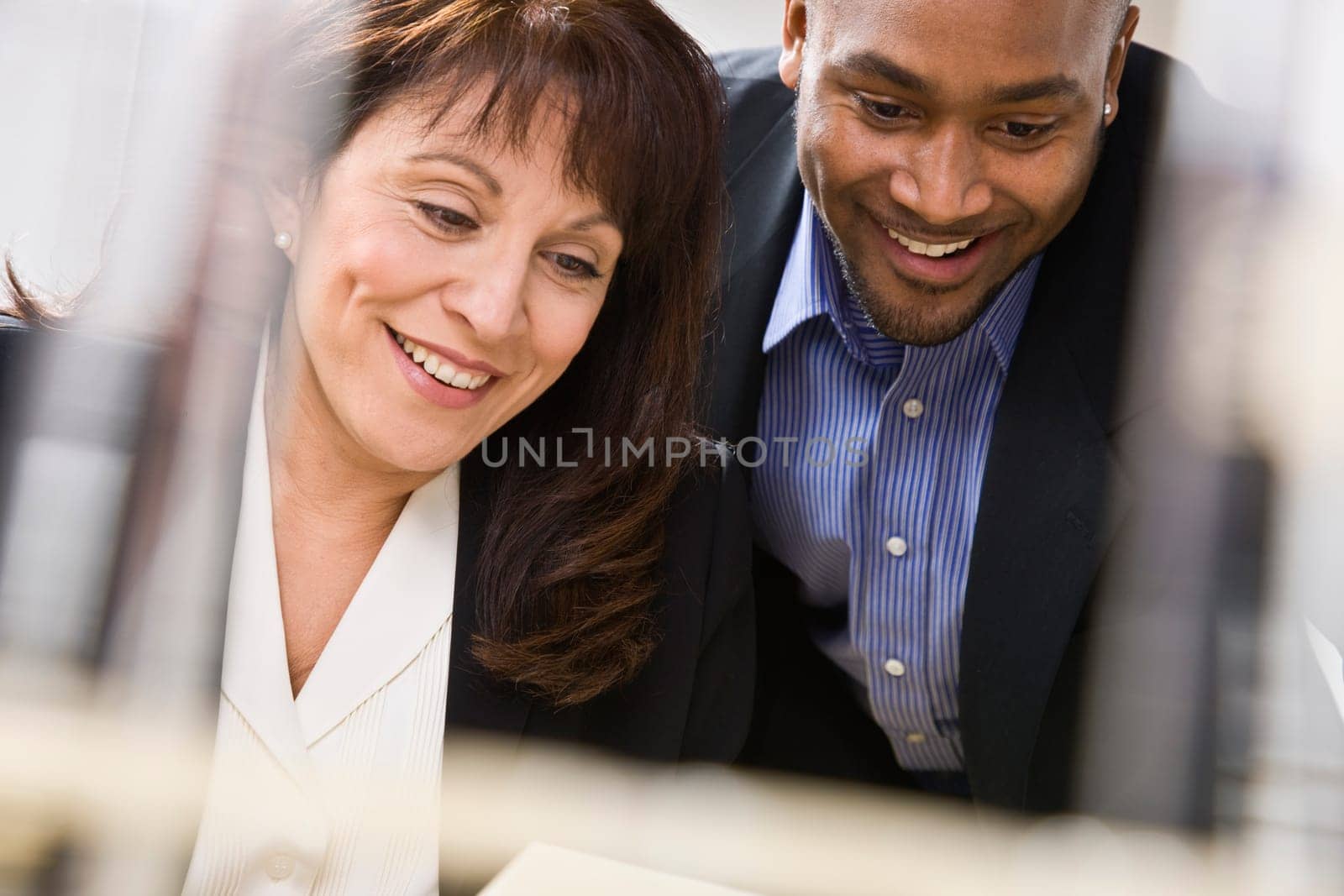A businessman and woman are working together in an office.  They are smiling and looking away from the camera.  Horizontally framed shot.