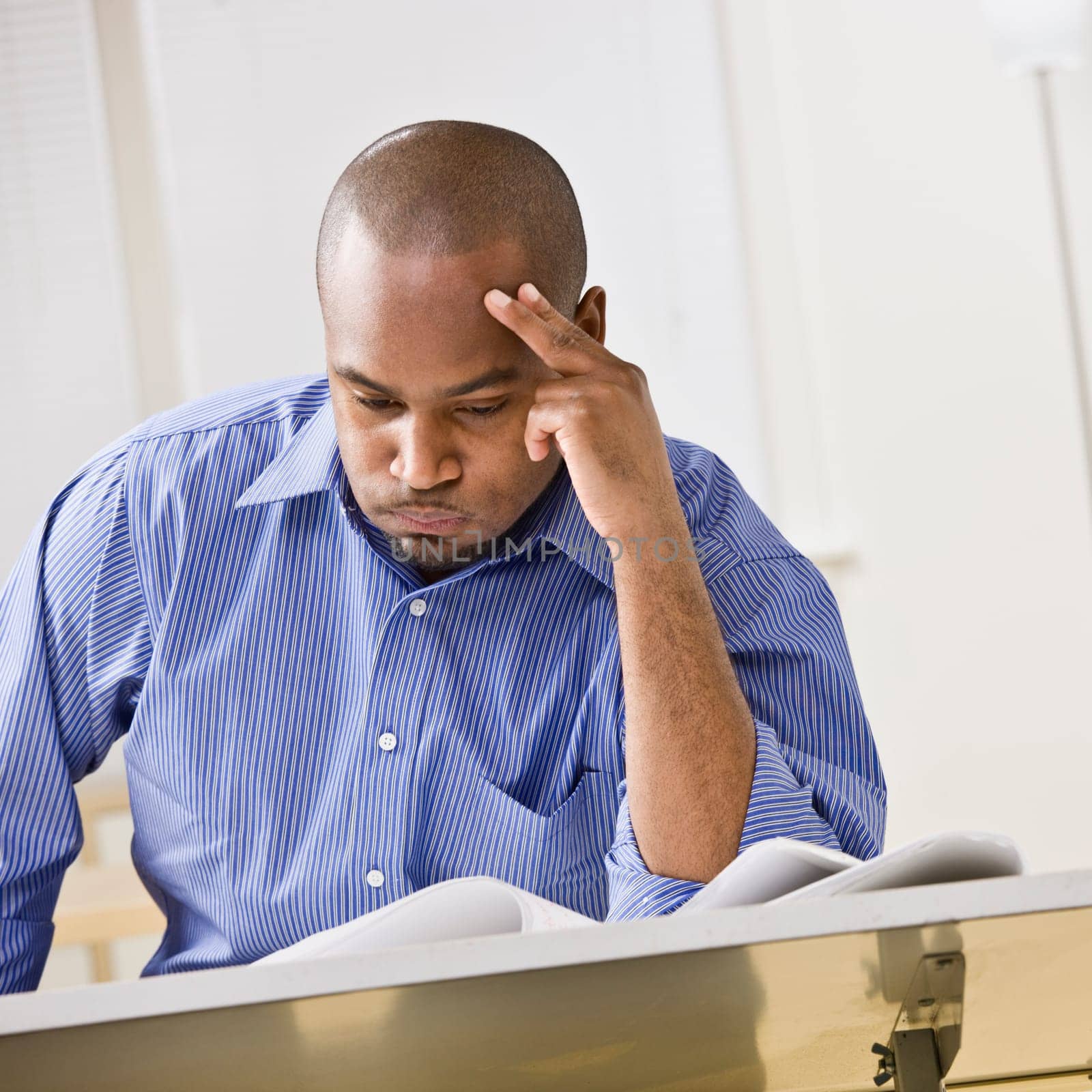 An attractive African-American male studying at a desk. He is resting his head against his fingers, and is puffing his cheeks out with air.  Square composition.