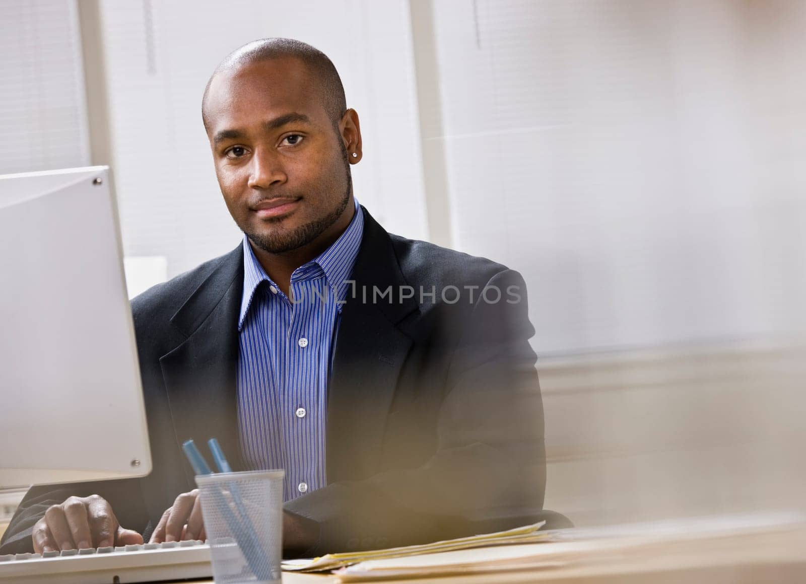 A young businessman is working on a computer in an office.  He is smiling at the camera.  Horizontally framed shot.