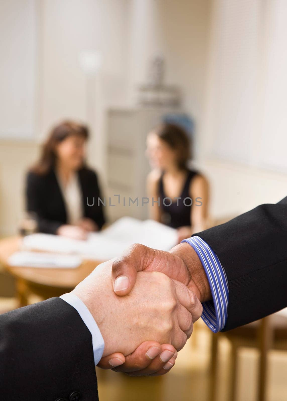 Two businesspeople are shaking hands in an office.  Two office ladies are seated at a desk in the background.  Vertically framed shot.